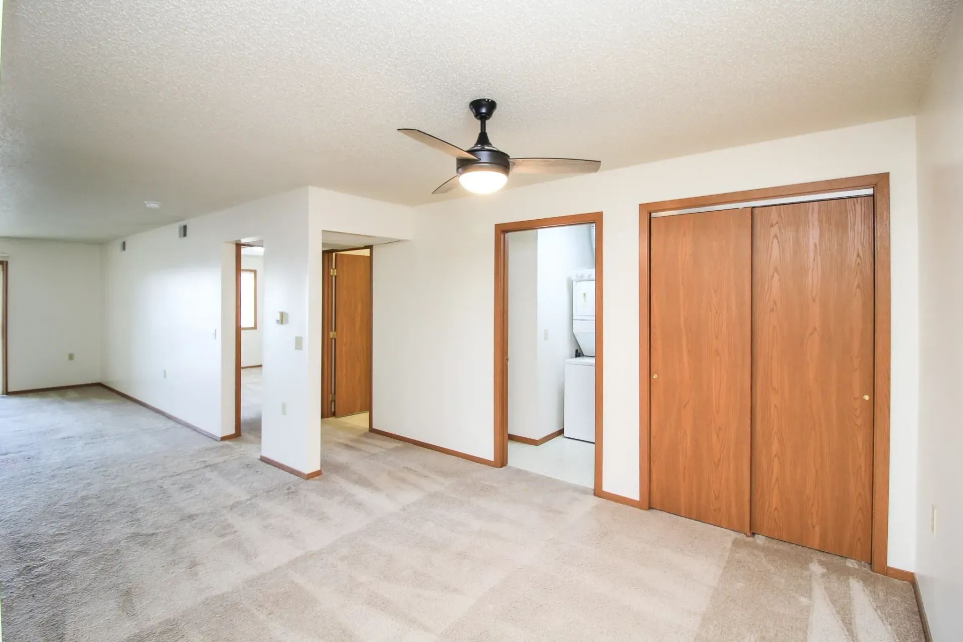 Empty beige bedroom with carpet, ceiling fan, white walls, and wooden closet doors
