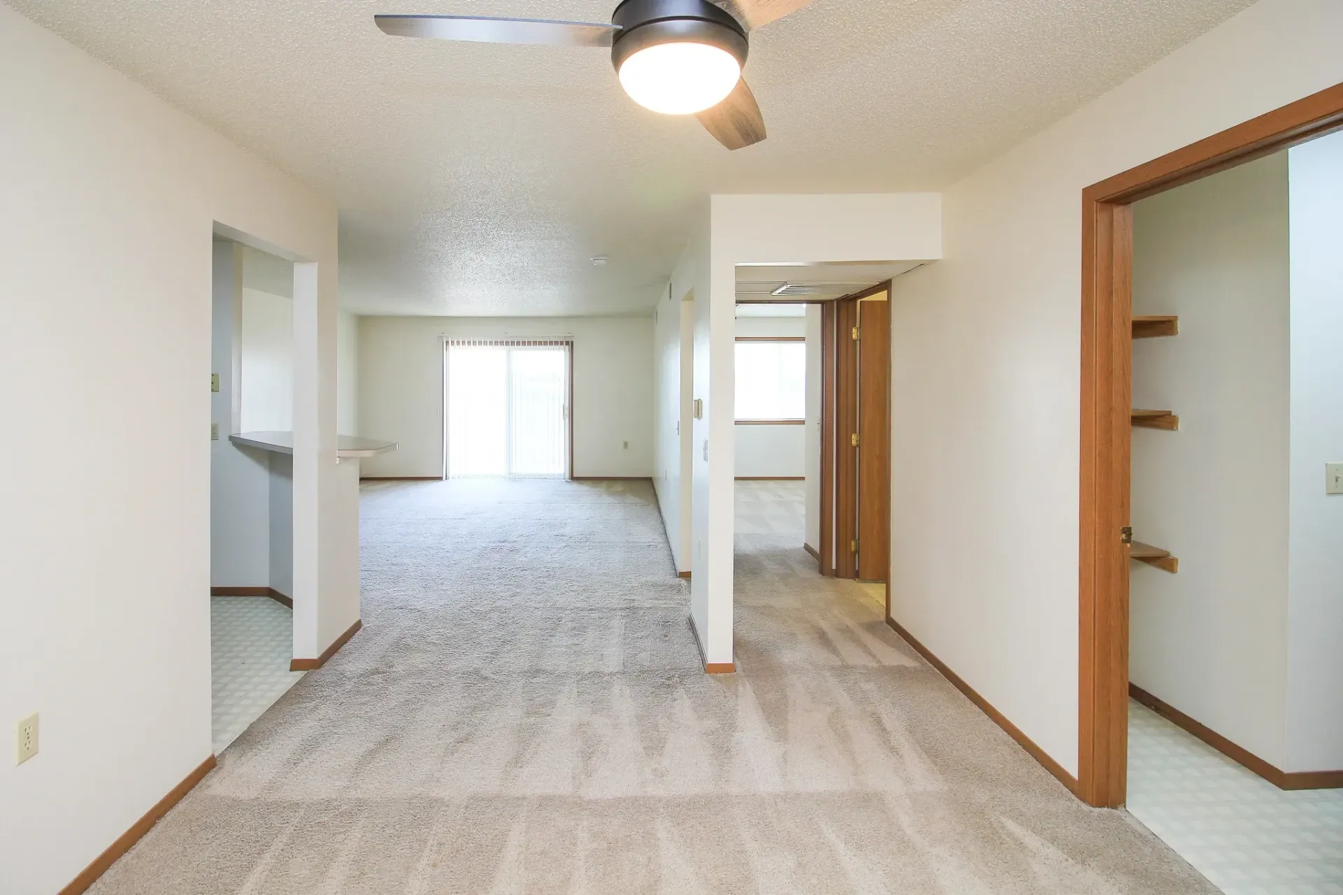 Empty carpeted apartment interior with white walls, ceiling fan, and wood-trimmed doorways