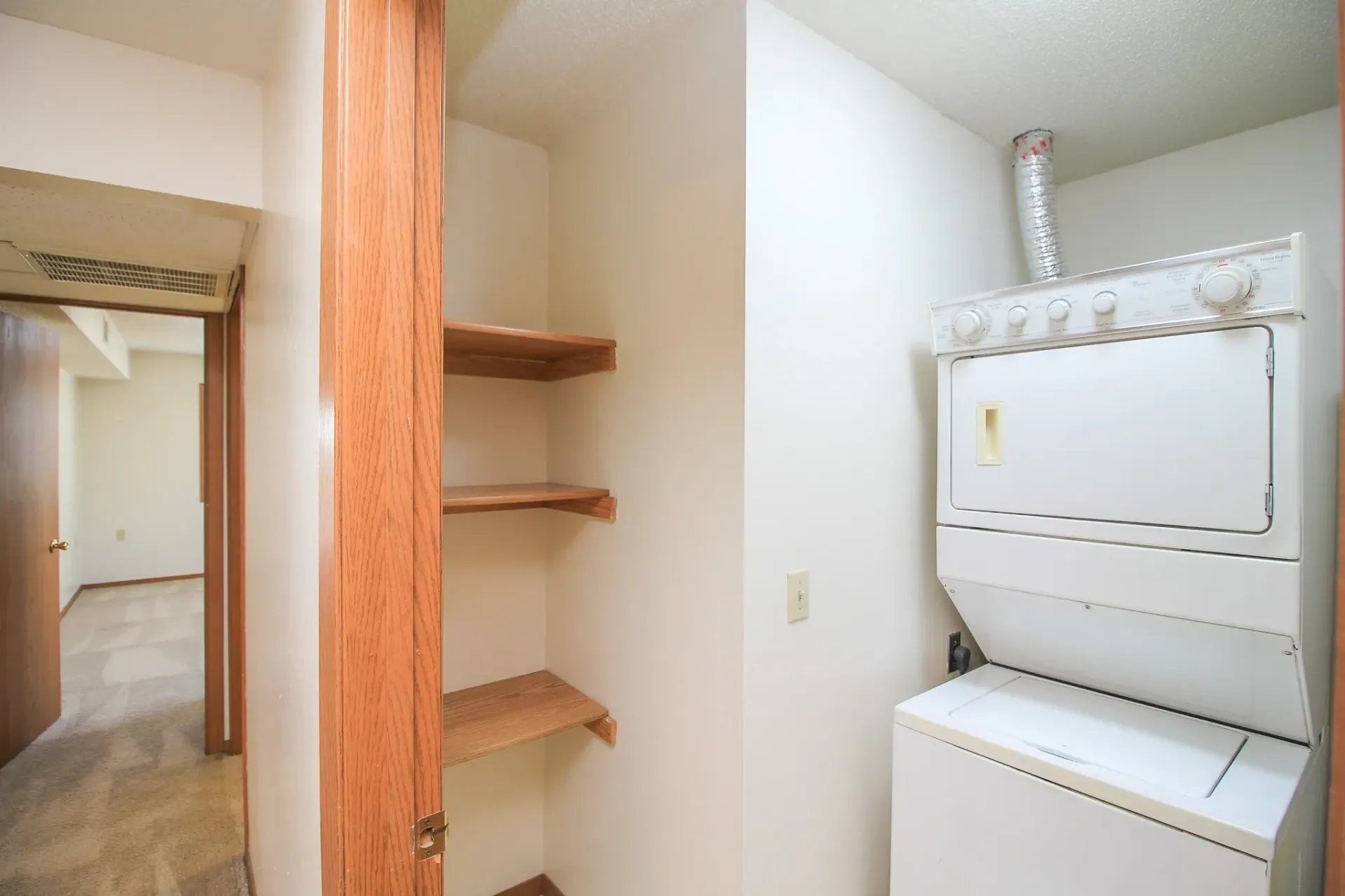 Laundry area with stacked white washer and dryer beside built-in wooden shelves and a hallway.