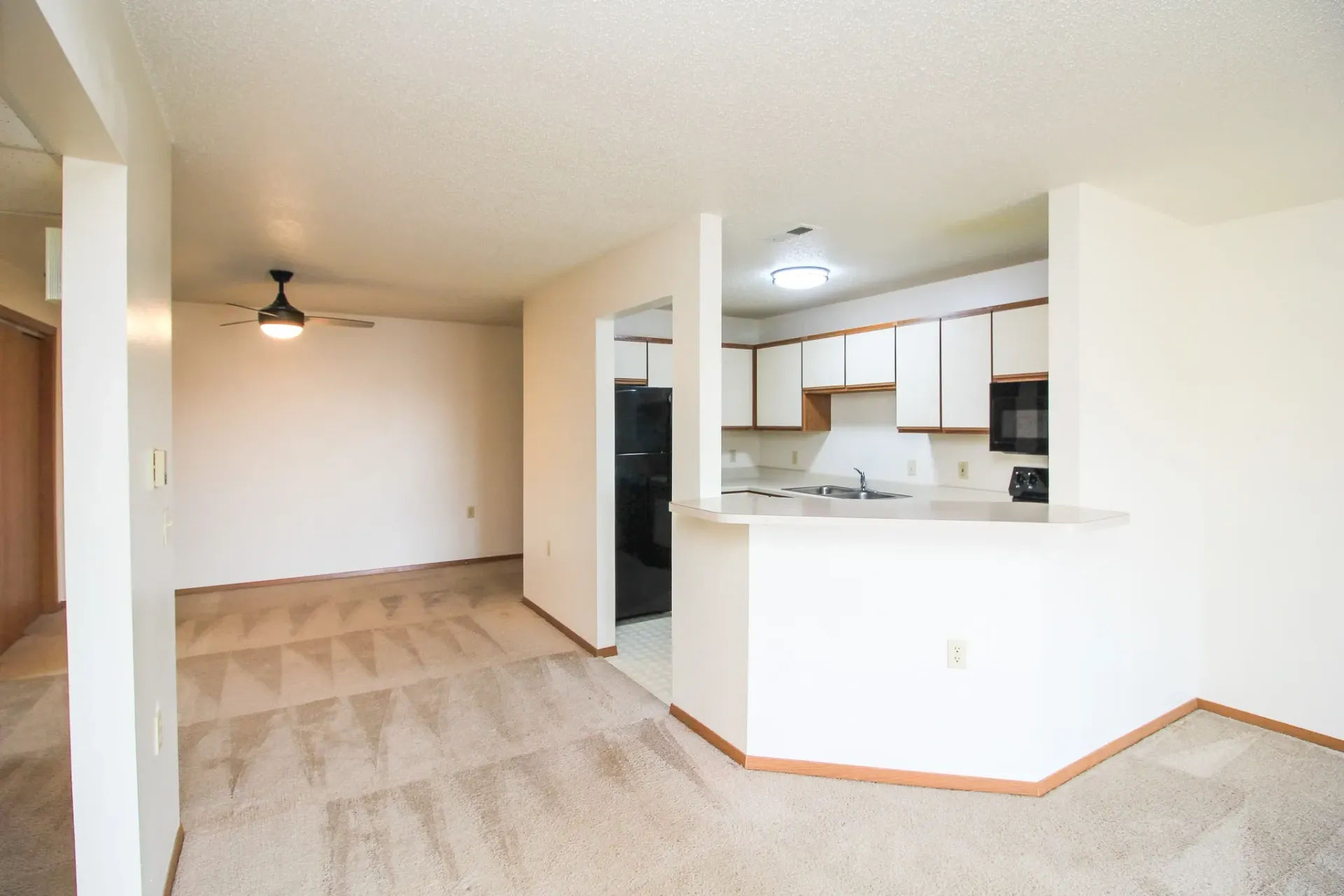 Empty living room with beige carpet, white walls, and a view into a kitchen pass-through.