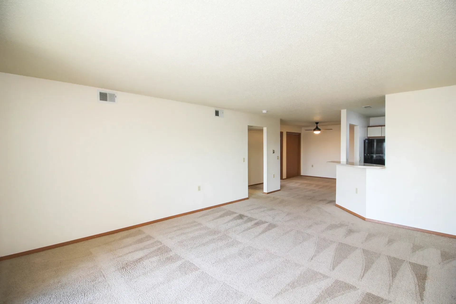 Empty beige carpeted living room with white walls and an open kitchen area