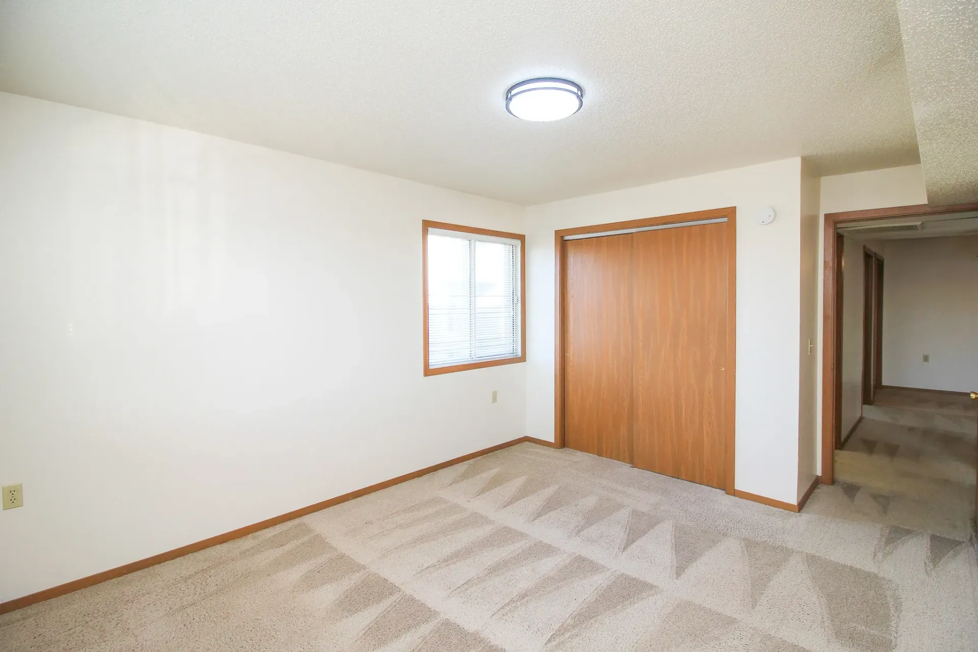 Empty carpeted room with white walls, a ceiling light, a window, and a wood closet door.