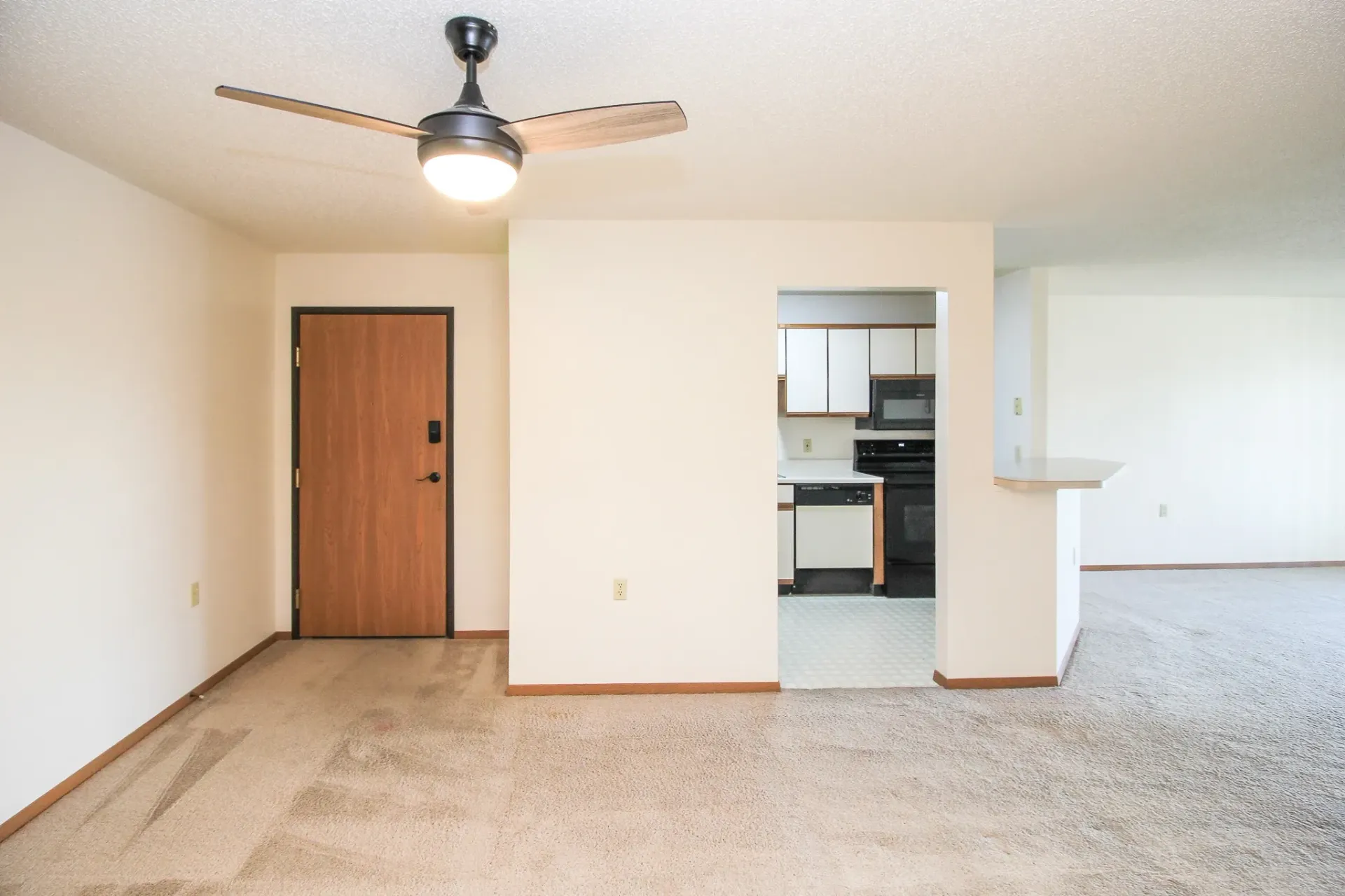 Empty carpeted living room with ceiling fan, wood door, and kitchen opening.