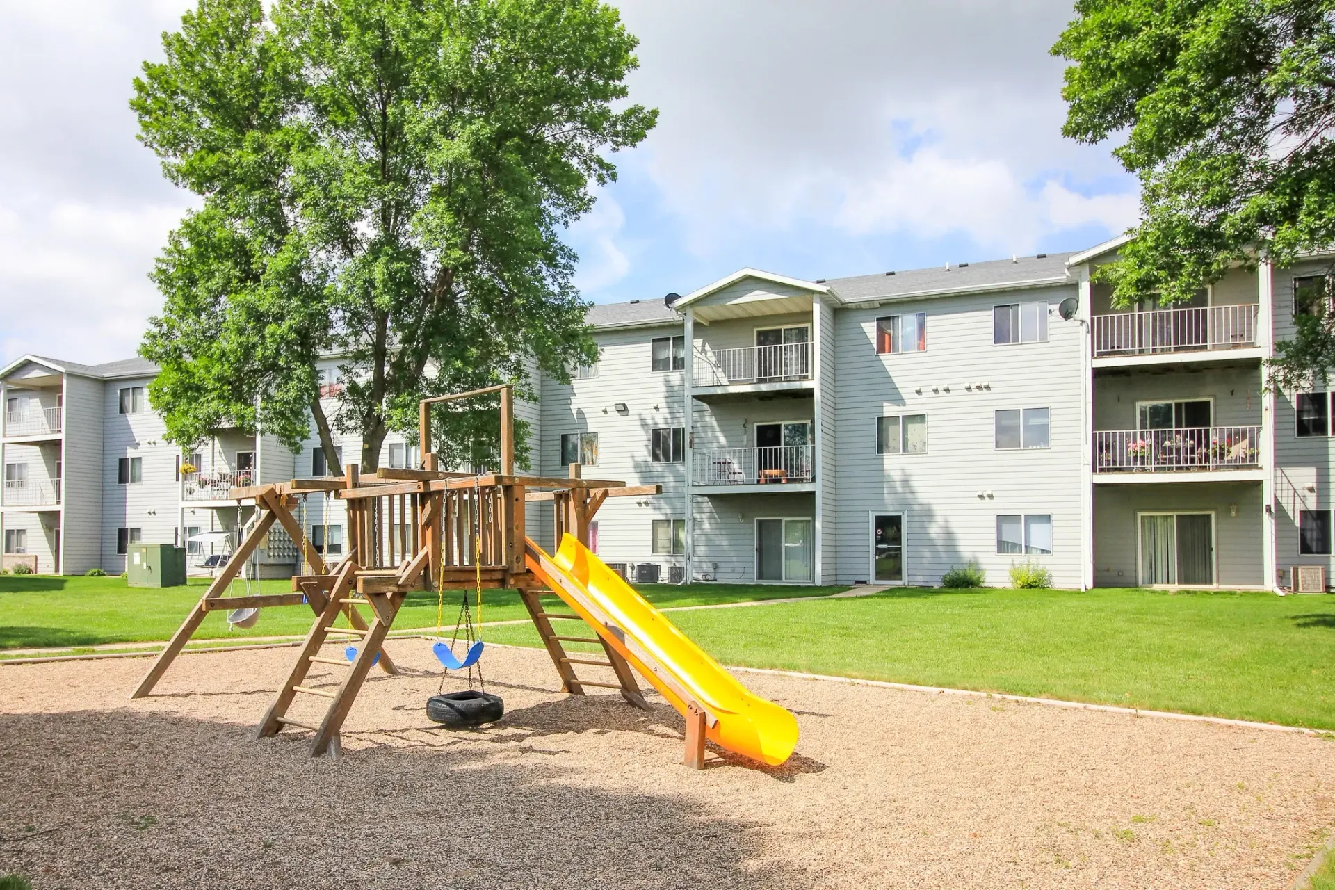 Playground with slide and wooden climbing frame in front of white apartment buildings and trees