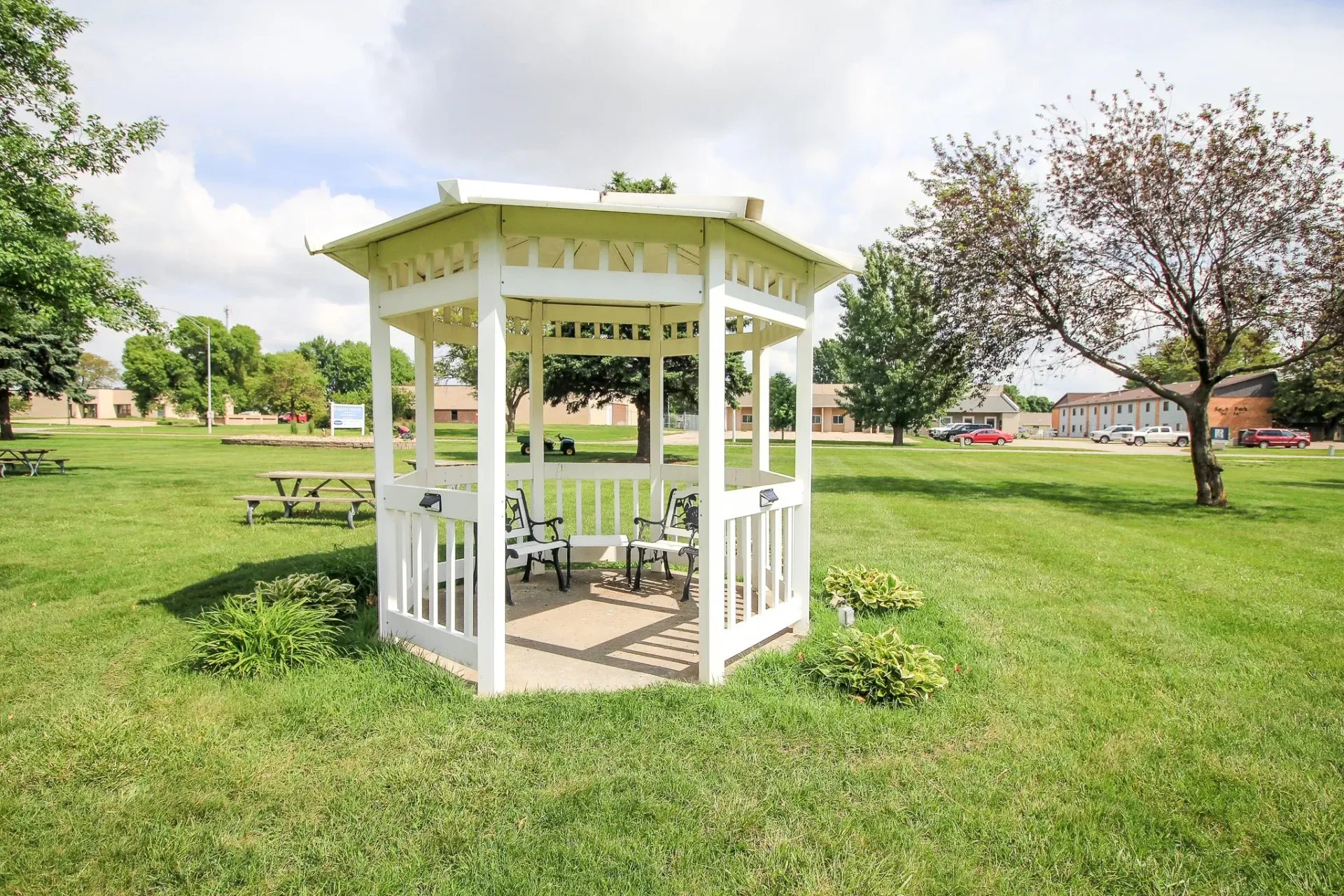 White garden gazebo on a grassy lawn under a cloudy sky, with trees and homes in the background
