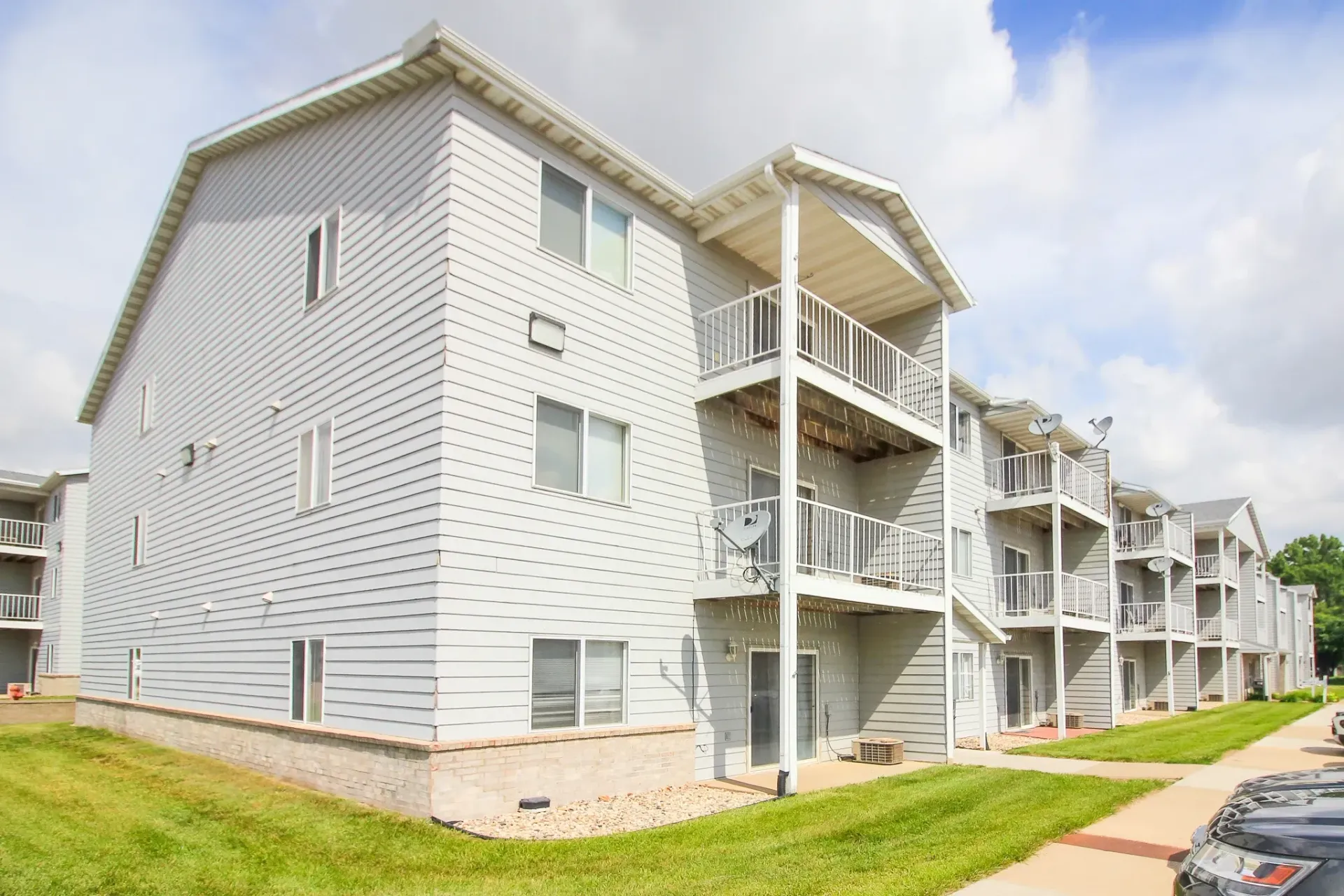 Modern gray apartment building with white balconies, spiral wall pattern, and a sidewalk beside green grass