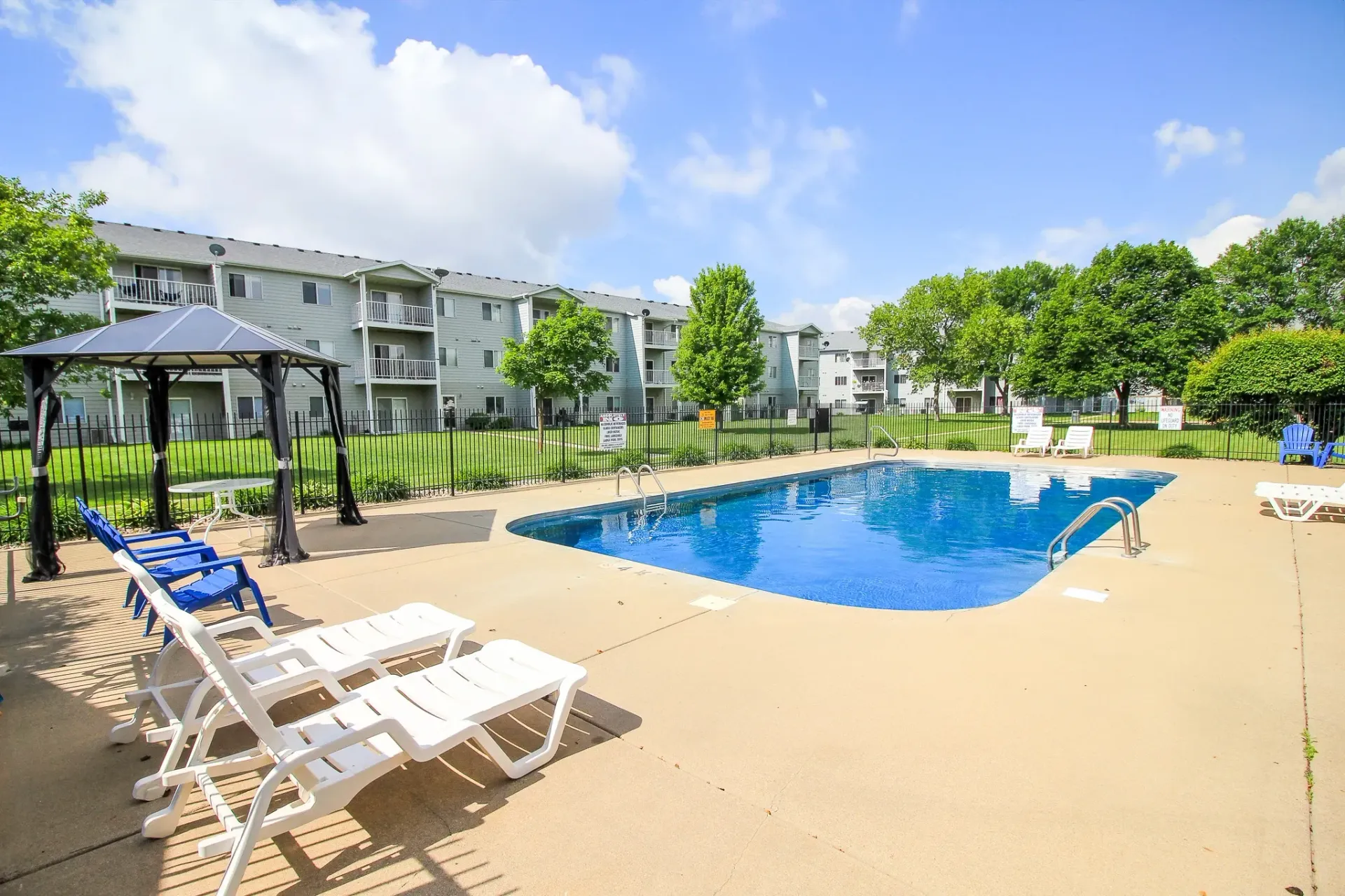 Apartment pool with lounge chairs, blue water, and trees under a bright sky