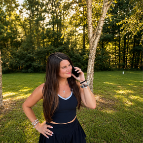 Woman in navy outfit smiling in a sunny park, holding a phone by a tree.