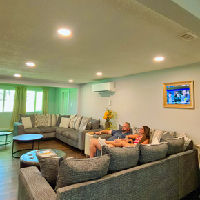 Bright living room with gray sectional sofas, two people relaxing, and a wall-mounted TV.