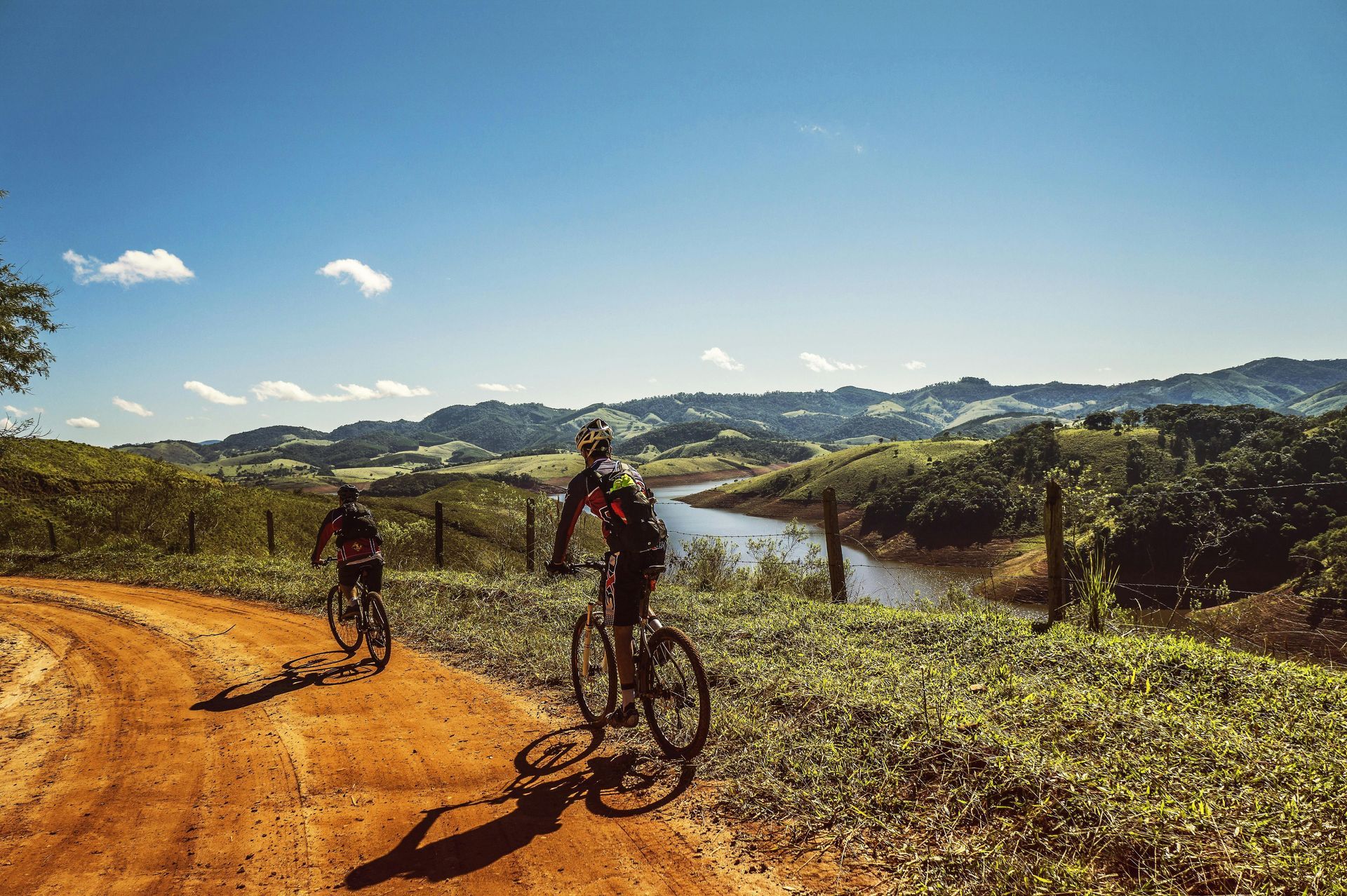 Cyclists on a dirt road overlooking green hills and a river under a bright blue sky