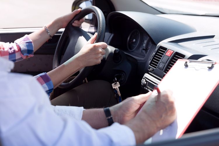 Driver's hands on steering wheel, receiving instructions from person with clipboard in car.
