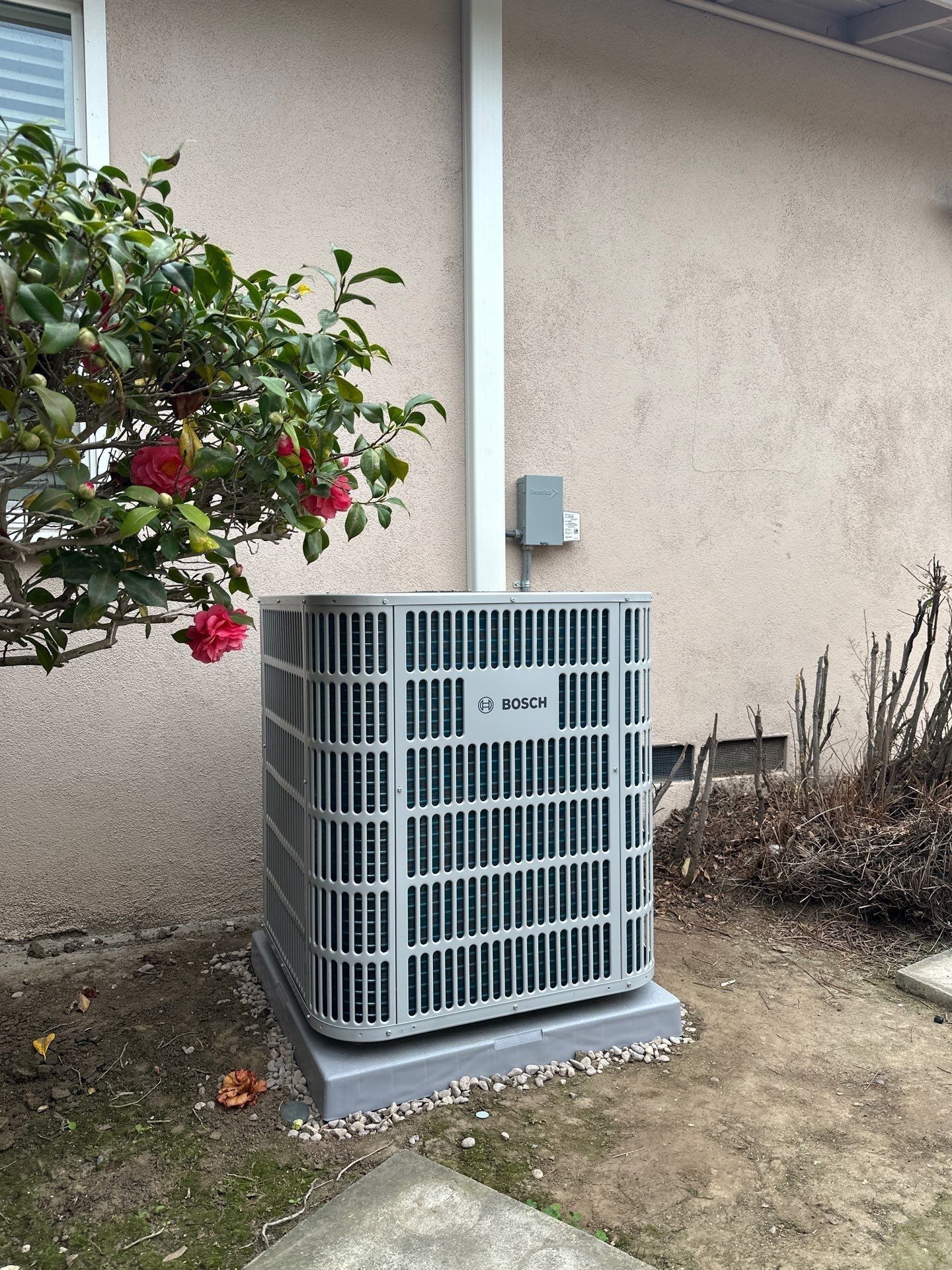 Air conditioning unit against a beige building, near a bush with red flowers, and a white pipe.