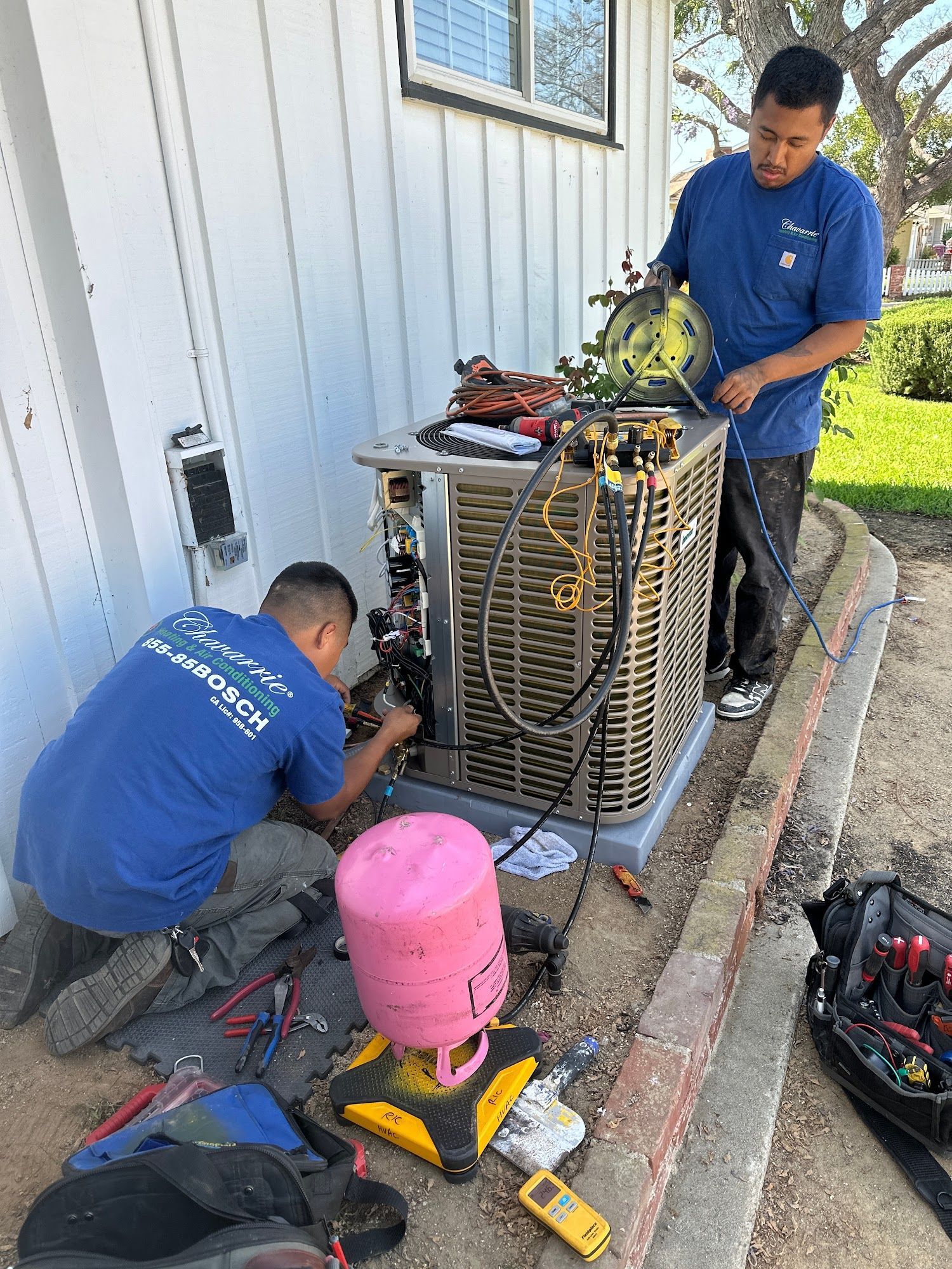 Two technicians in blue shirts repairing an AC unit outside a house; various tools are present.