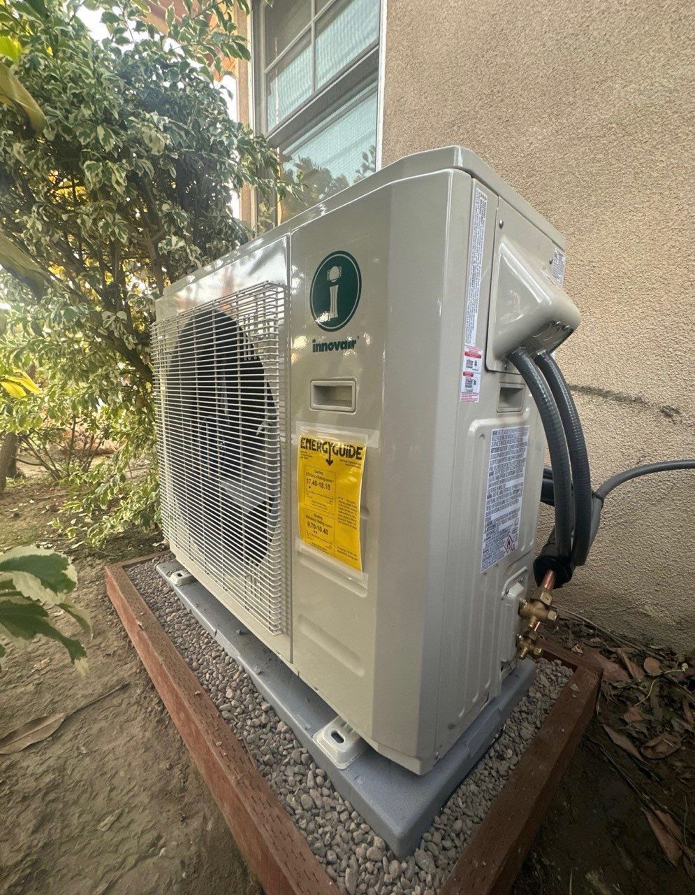 An outdoor heat pump unit, beige, mounted on a concrete base, next to a building and greenery.