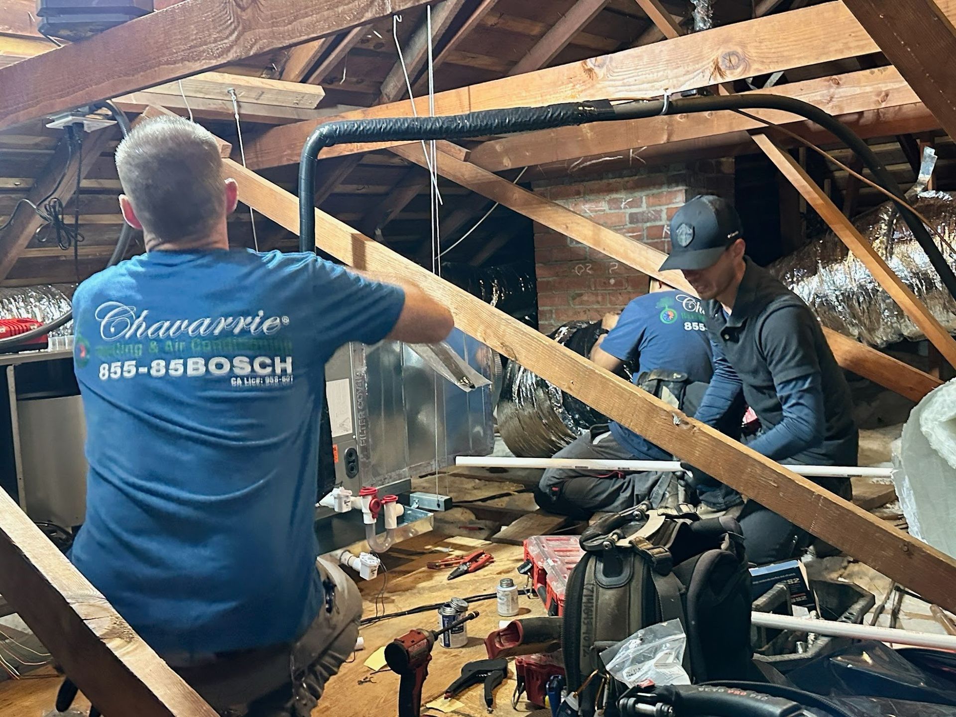Workers installing HVAC system in an attic. One man in blue shirt, other in black cap.
