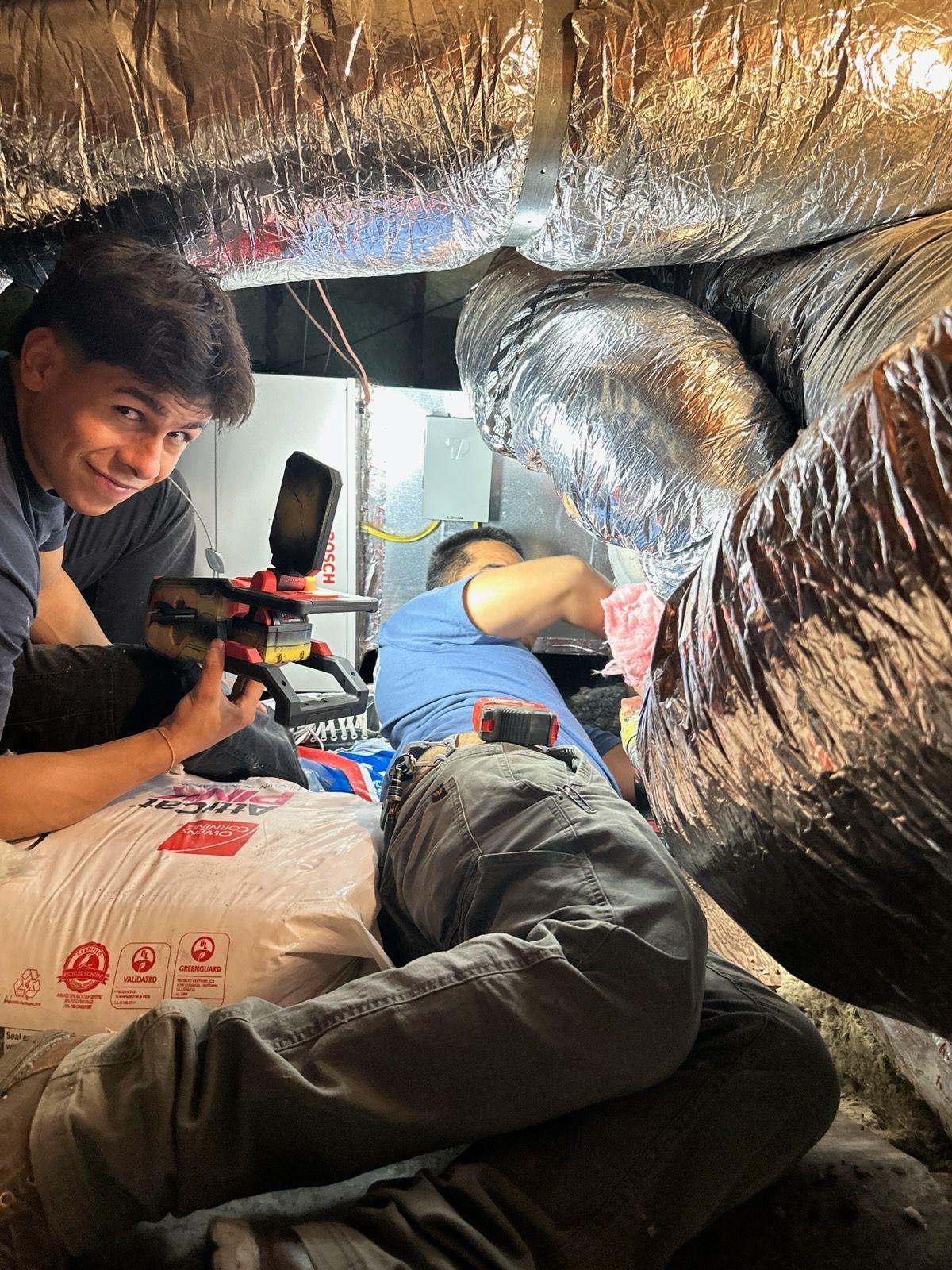 Two people work on HVAC system in a crawl space. One holds a light, the other works on equipment.