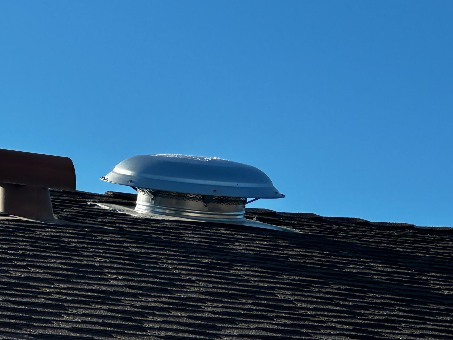 A metal roof vent on a dark shingled roof against a bright blue sky.
