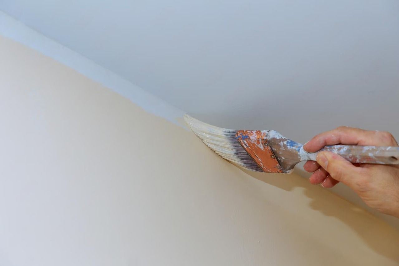 Hand painting the edge of a ceiling with a brush, applying white paint to a beige wall