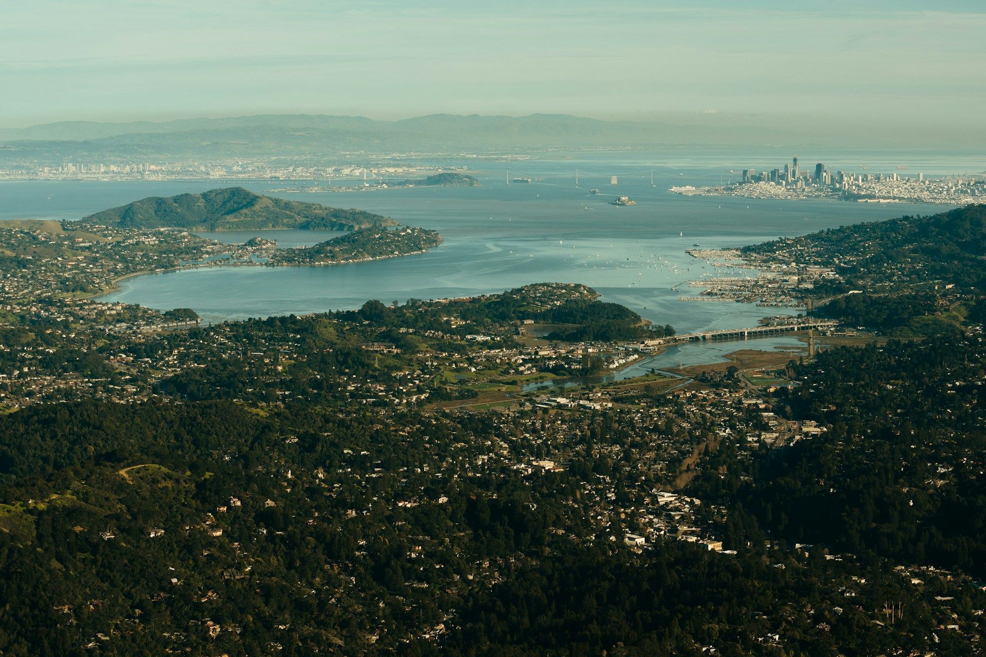 Aerial view of a bay and city skyline beyond forested hills under a hazy sky