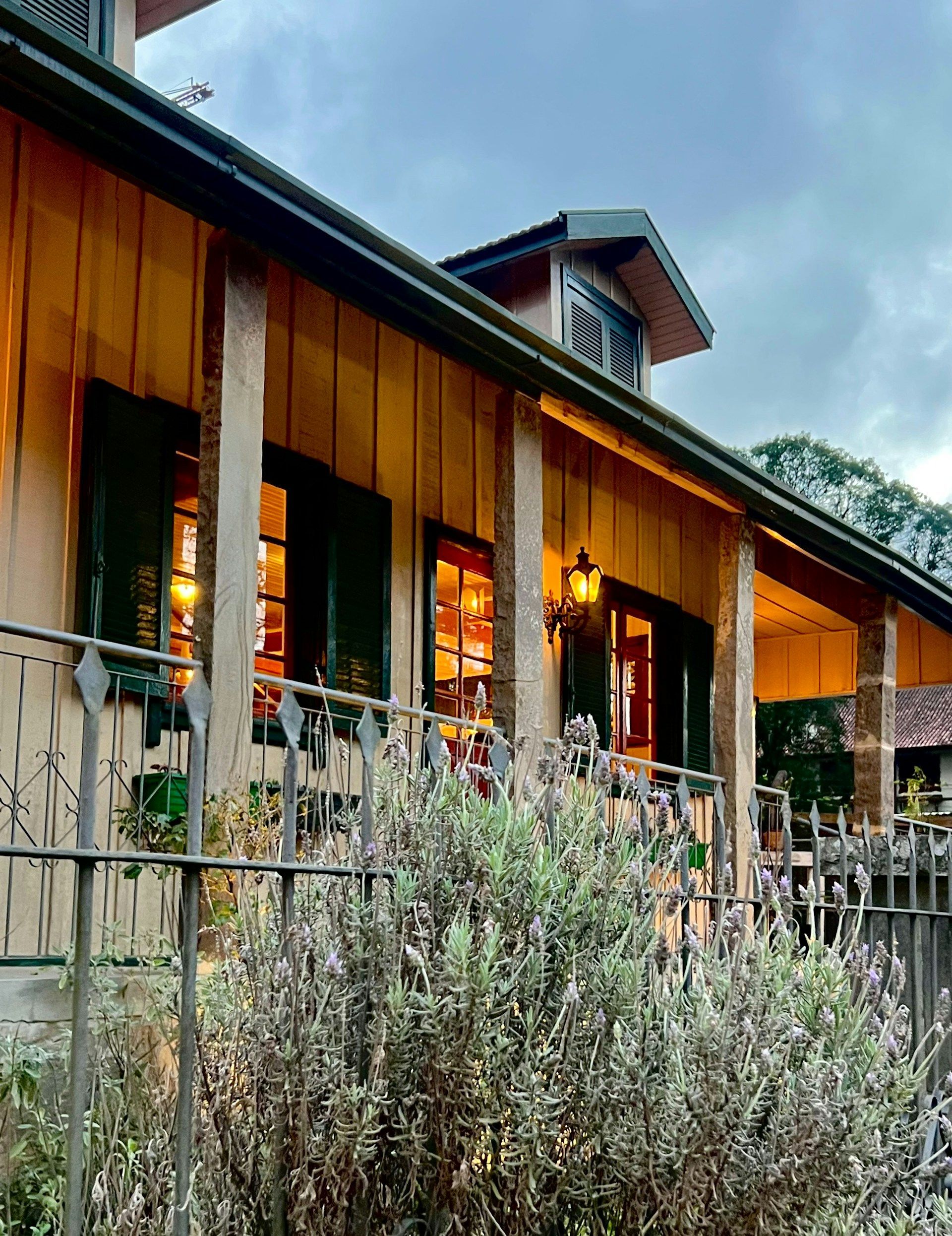 Wooden house with warm-lit windows behind a rustic fence and shrubs at dusk