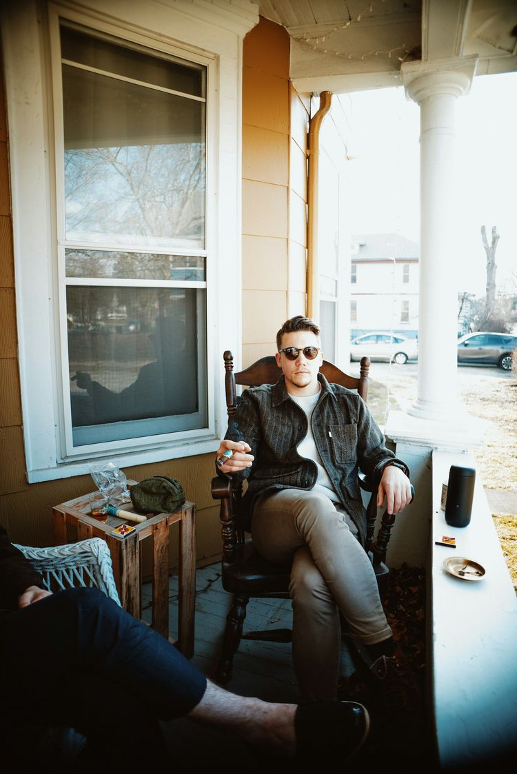 Person in sunglasses sits on a porch chair holding a drink, with another person’s legs in the foreground.