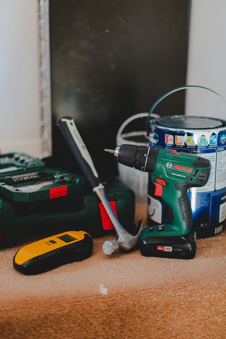 Tools on a carpeted floor: drill, hammer, tape measure, and paint bucket beside a wall.