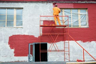 Worker on orange scaffold painting a building’s white brick wall red.