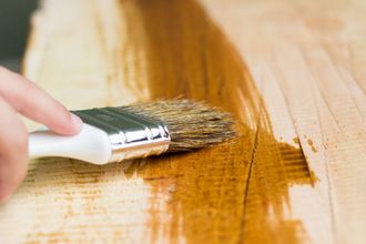 A hand painting a wooden surface with a white-handled brush, applying a glossy amber stain.