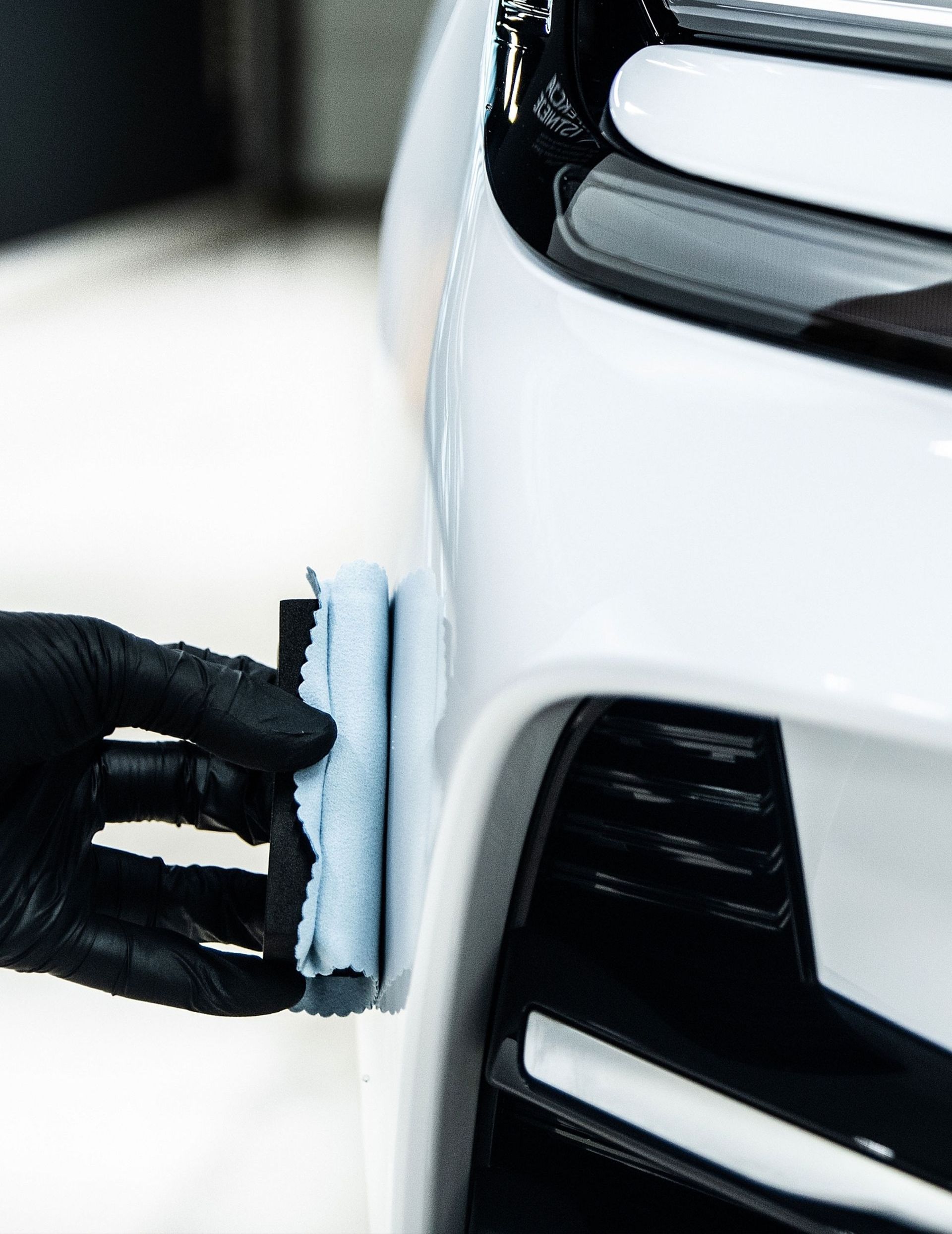 A person is cleaning the front of a white car with a sponge.