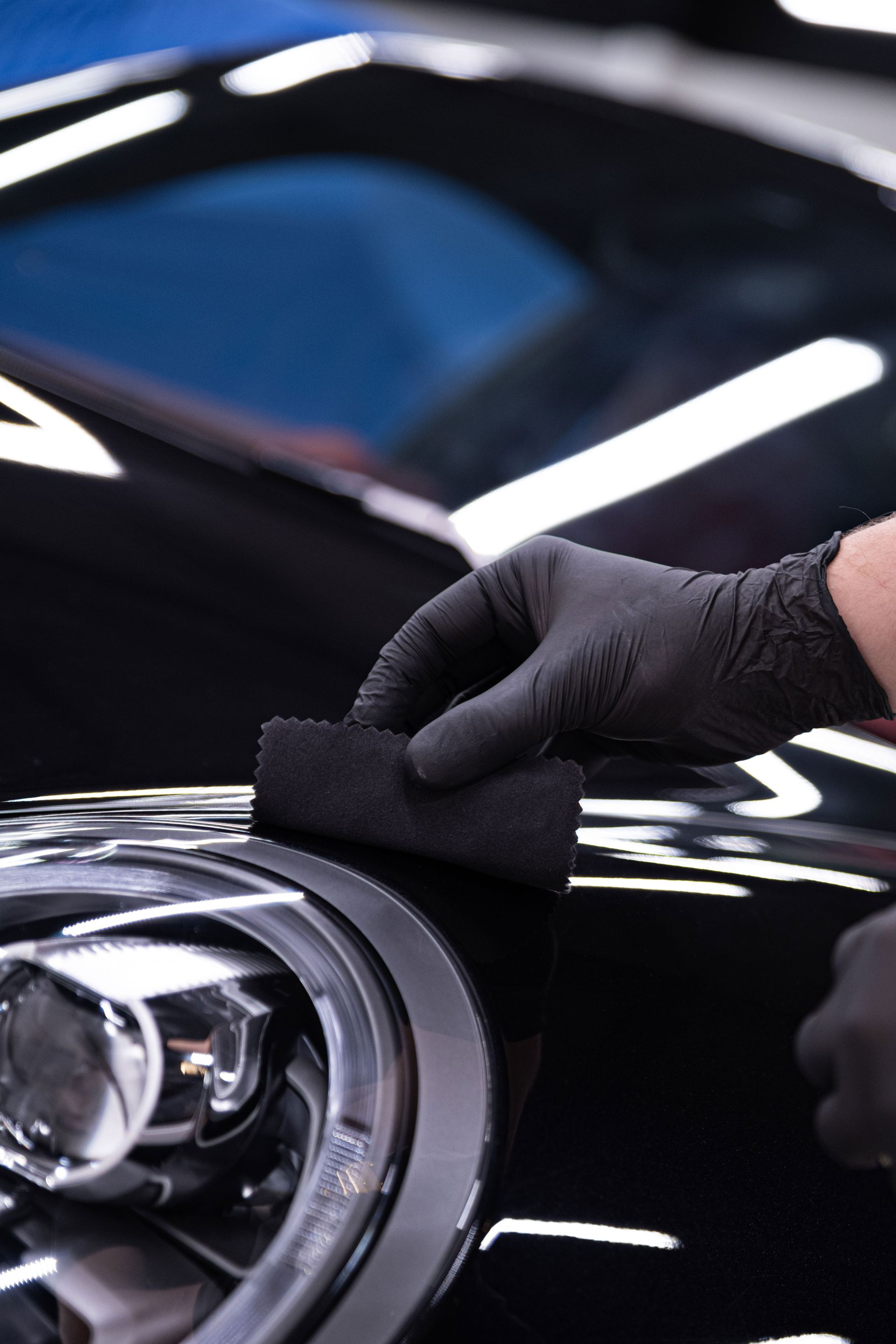 Person in black gloves applying a ceramic coating to a black car's headlight with a black applicator.