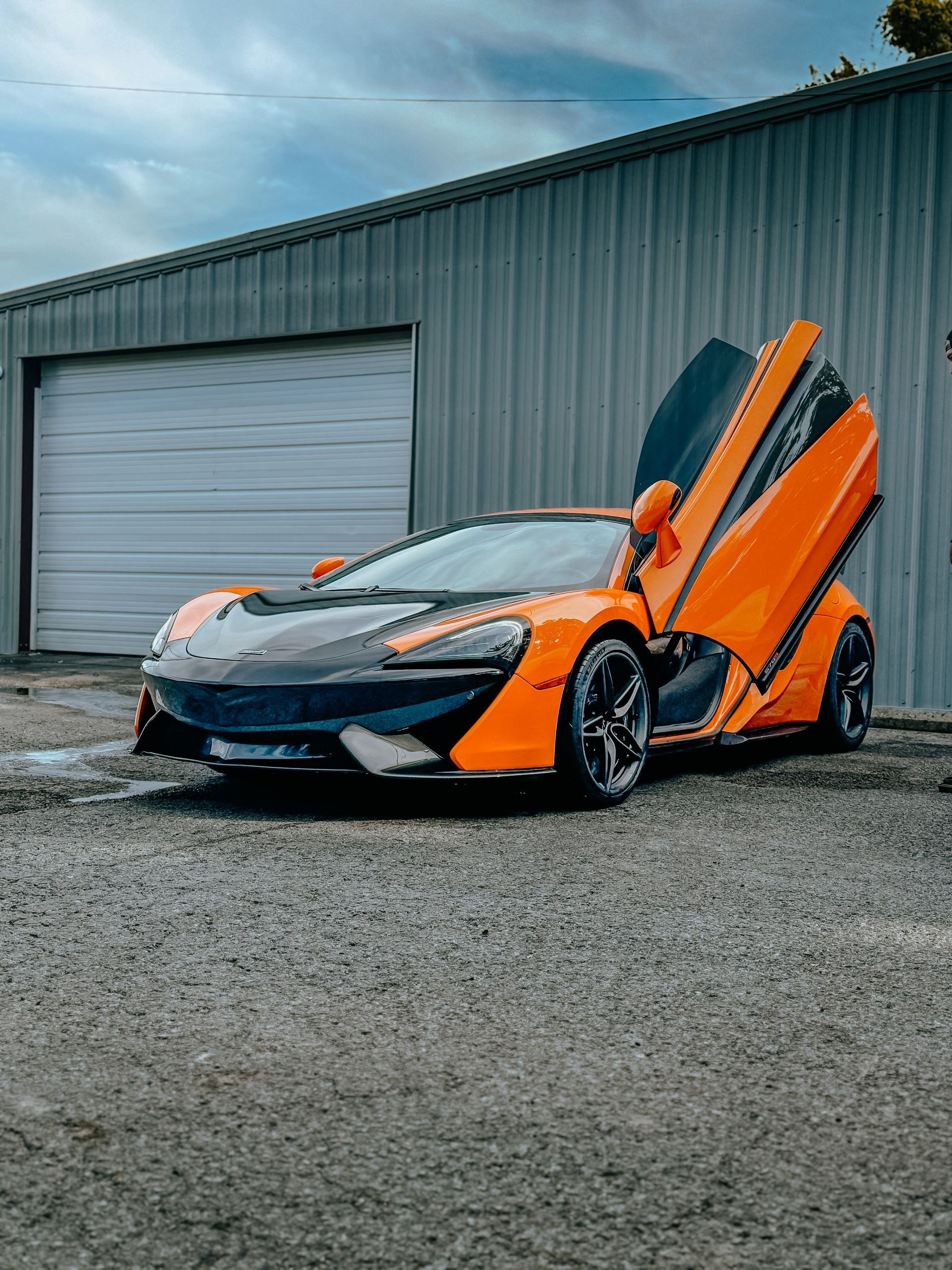 Orange sports car with butterfly doors open, parked outside a gray garage on a gravel lot