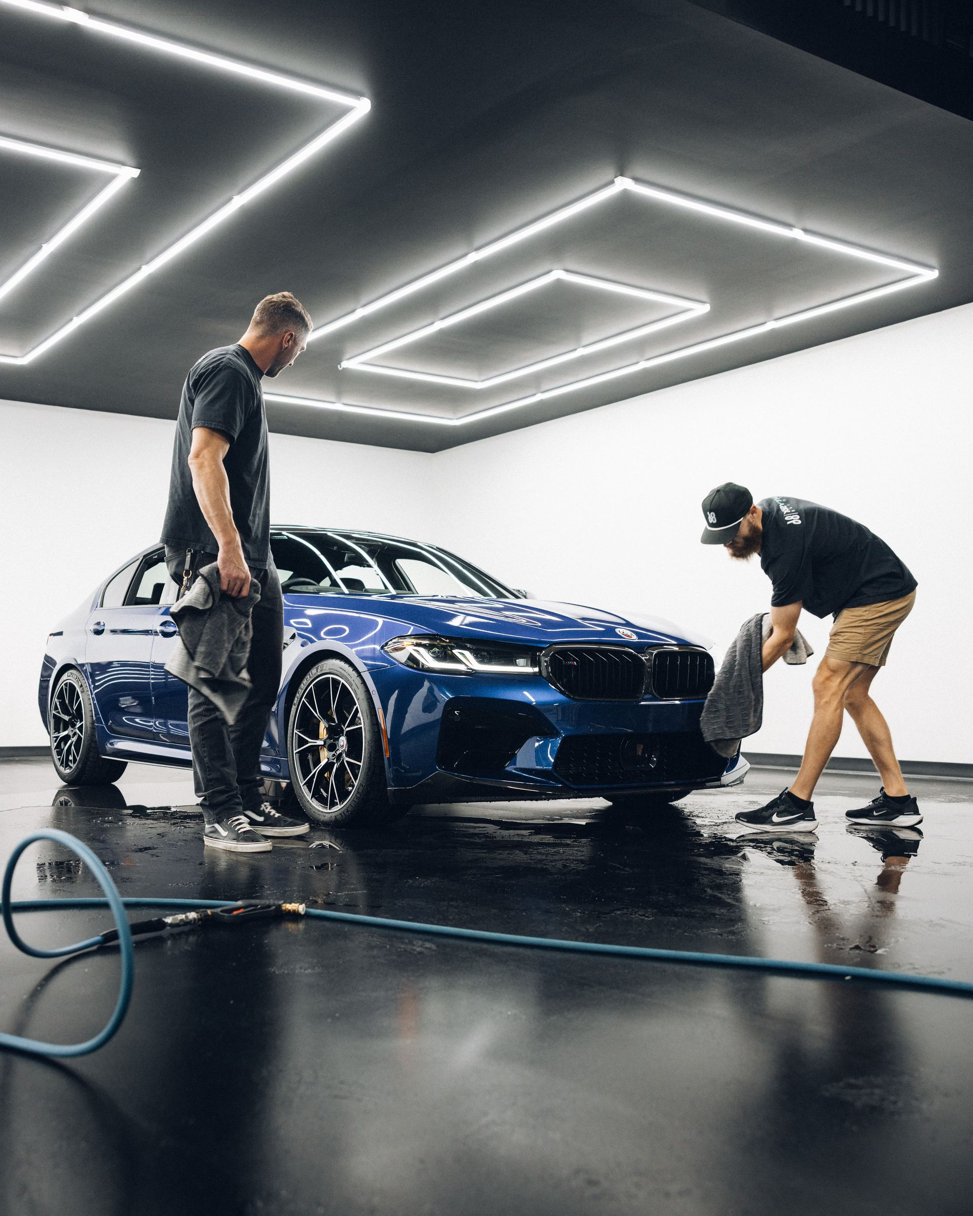 Two people cleaning a dark blue BMW in a well-lit garage, one drying the car, the other observing.