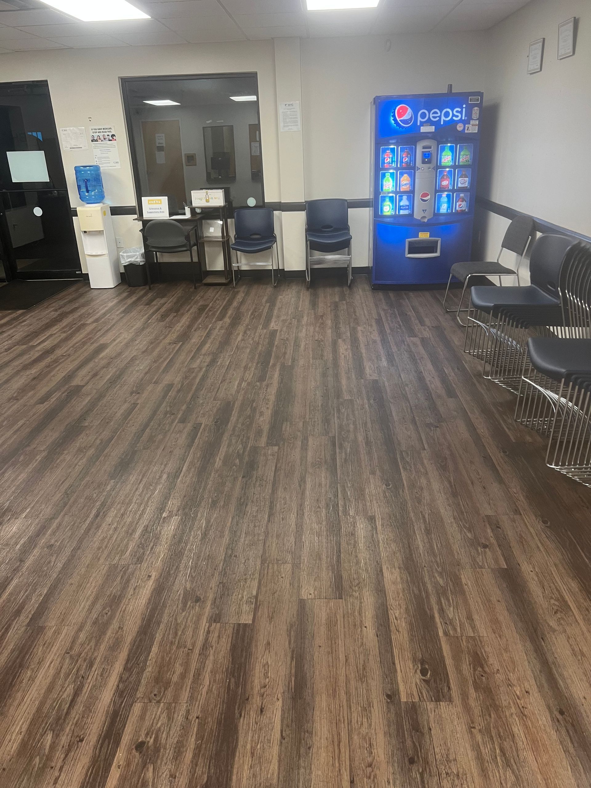 Empty lobby with wood-pattern floor, chairs along the walls, and a blue vending machine near the back.