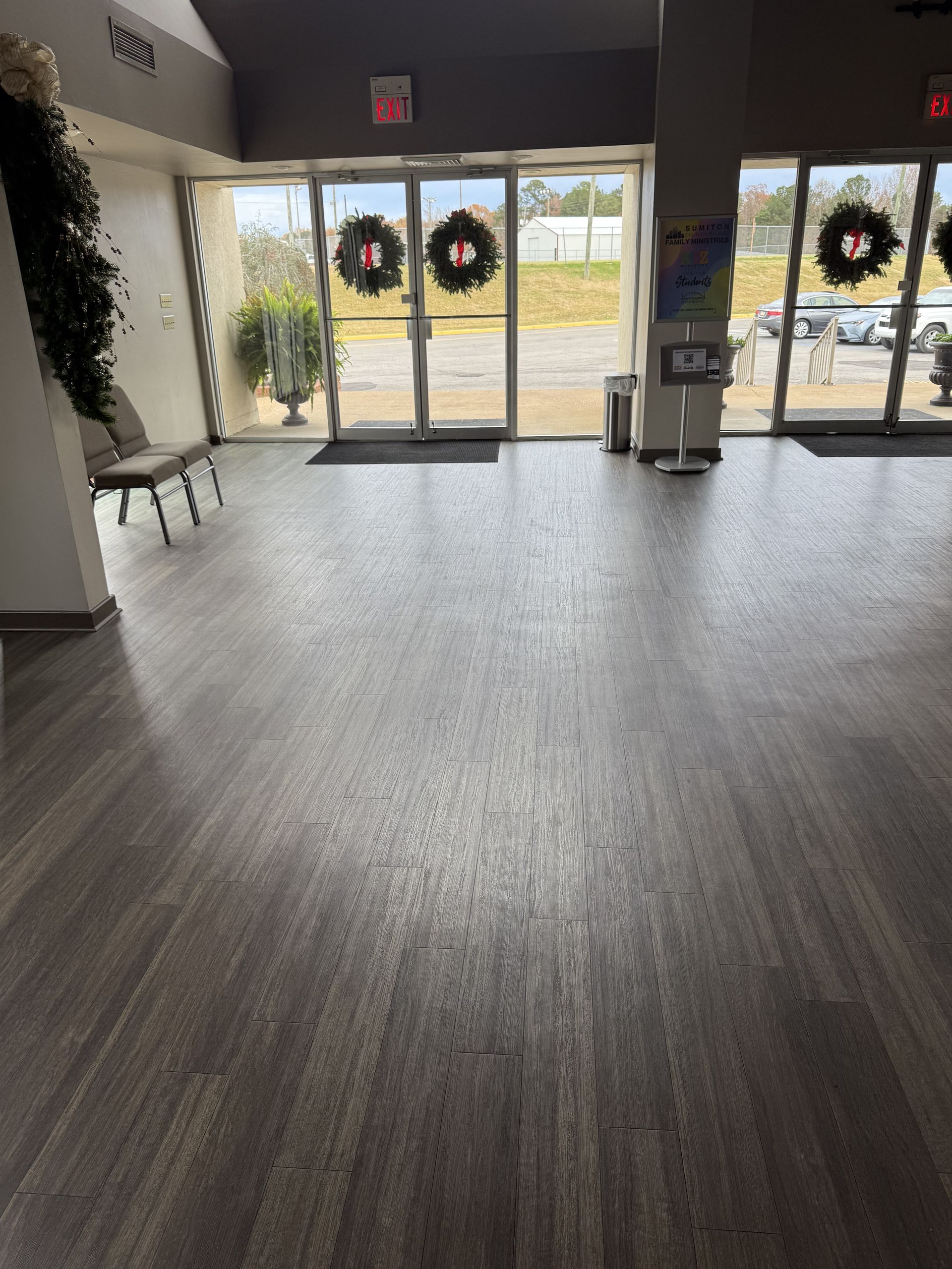 Empty lobby with glass doors decorated with wreaths, wood-look flooring, and a plant near the wall.