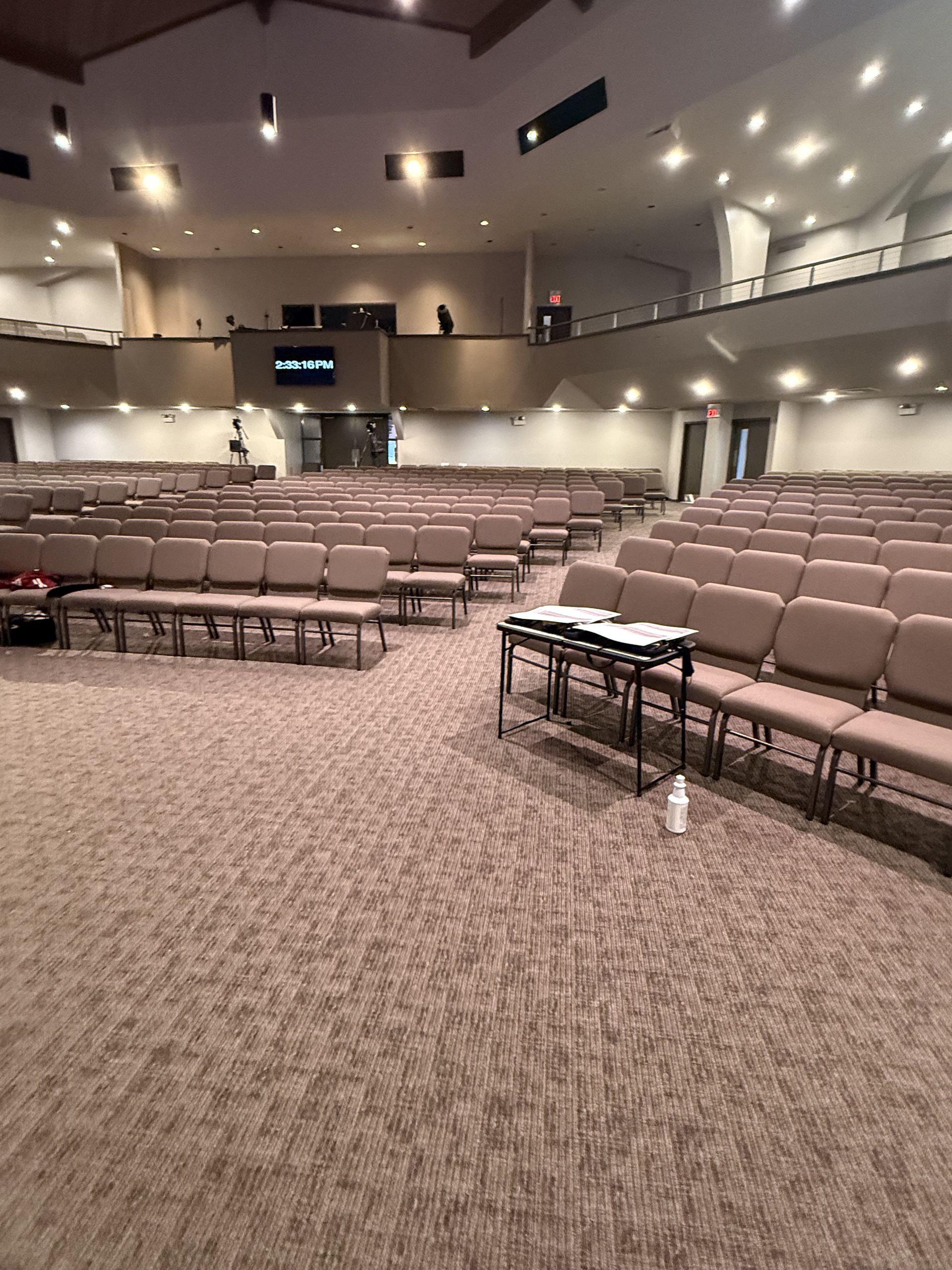 Empty auditorium with rows of beige seats and a carpeted stage area under recessed ceiling lights