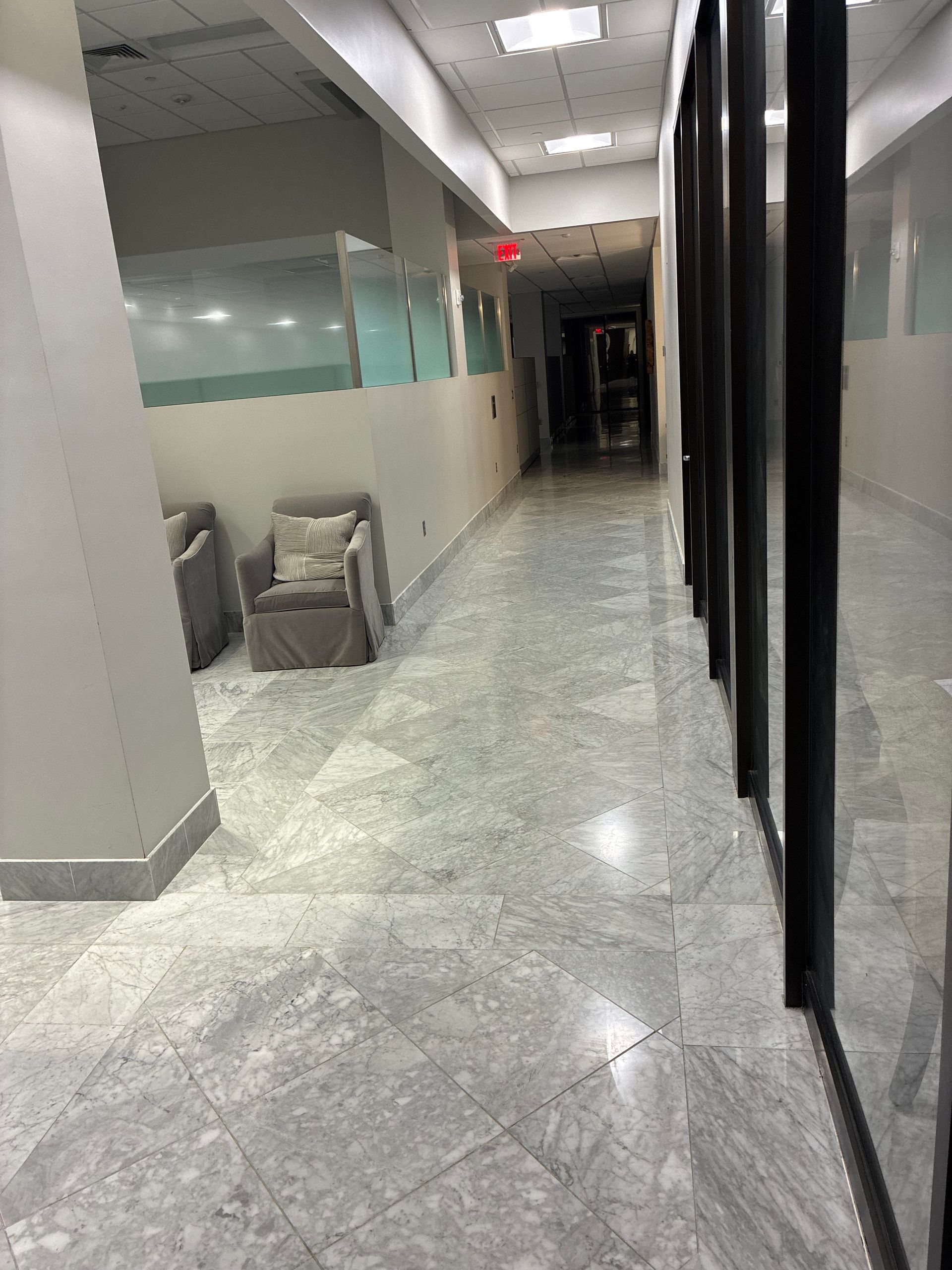 Empty office hallway with gray tiled floor, glass walls, and a chair near the left wall.