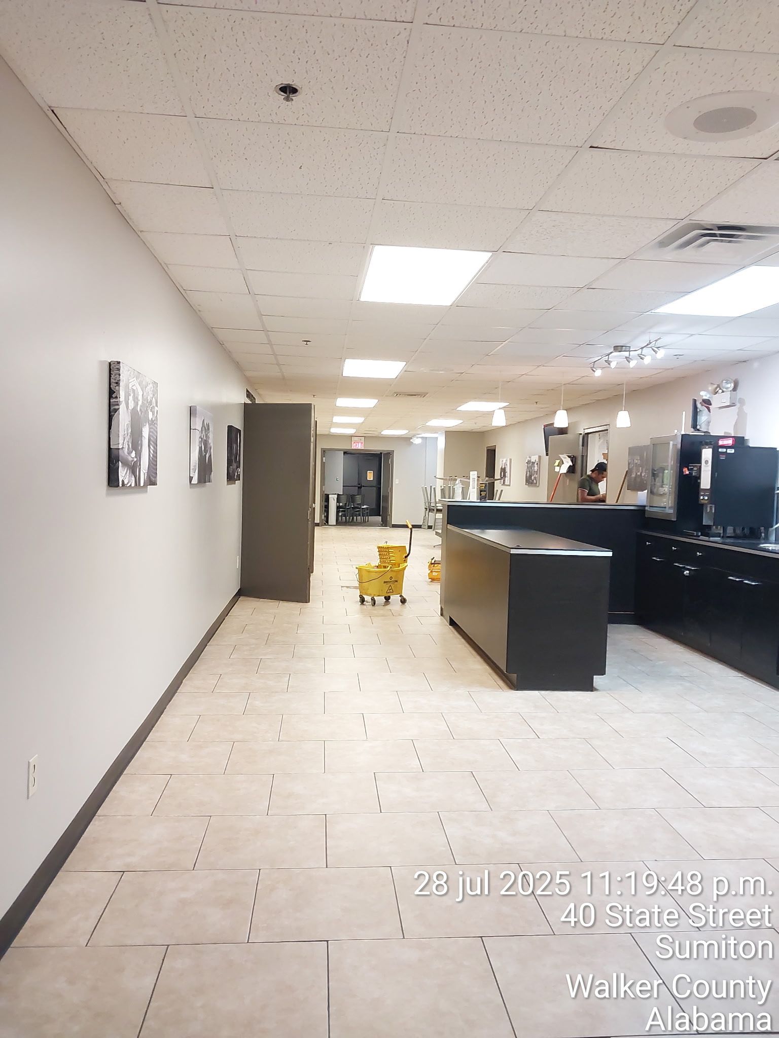 Empty office hallway with tiled floor, white walls, desk area, and a yellow wet floor sign under ceiling lights