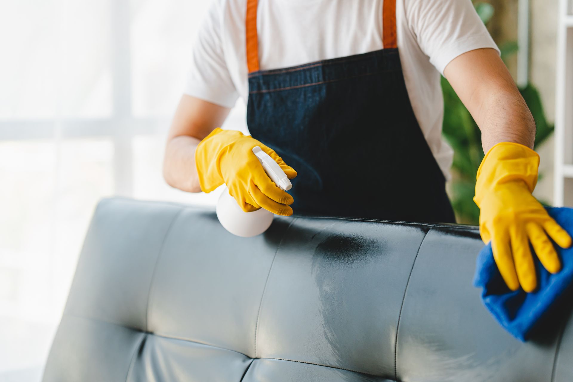 Person wearing yellow gloves cleaning a gray sofa with a blue cloth