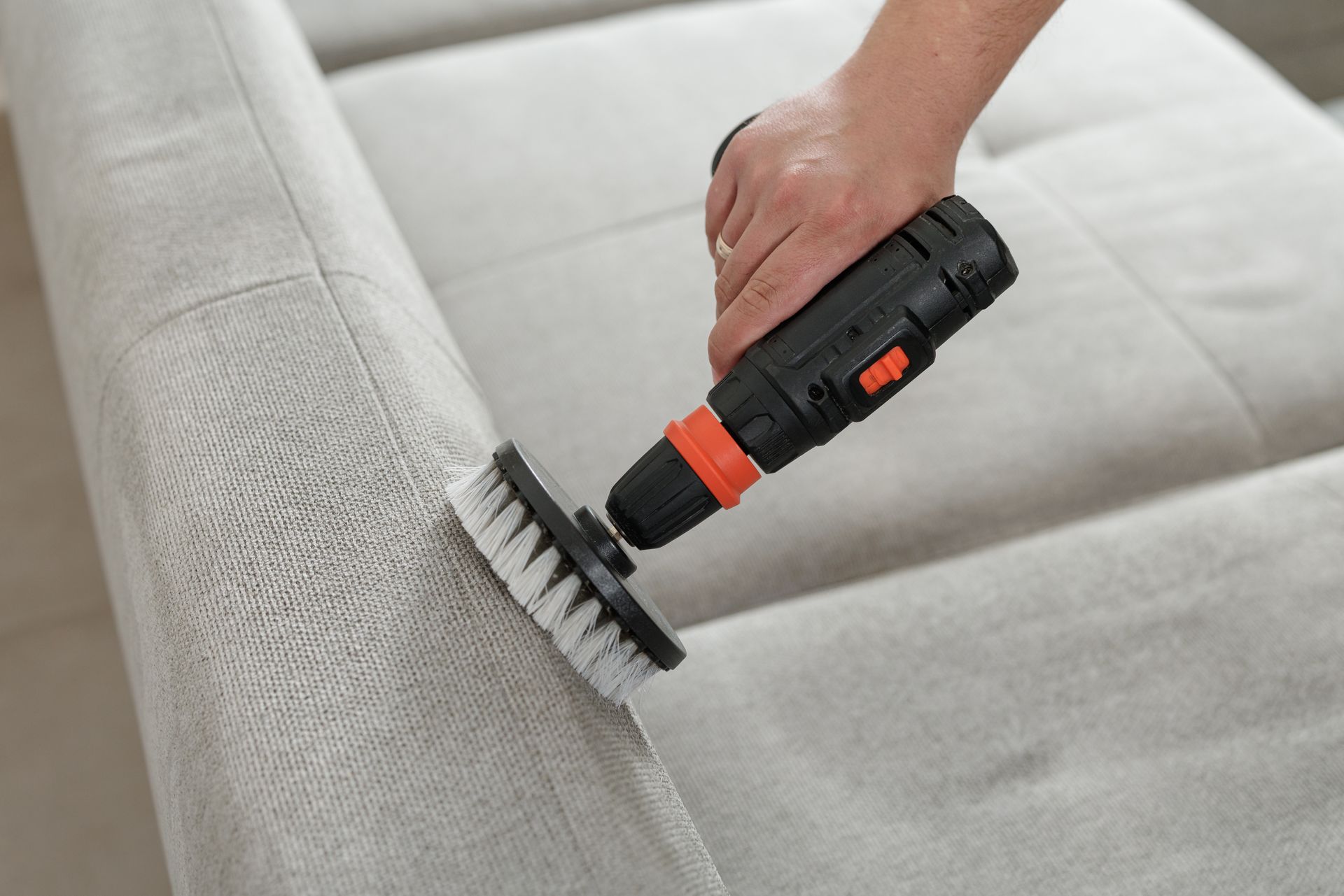 Hand using a black handheld vacuum with orange accents to clean a gray couch seam