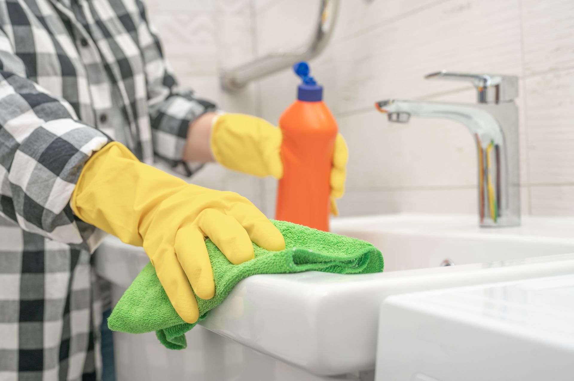 Gloved hands cleaning a white sink with a green sponge and orange spray bottle in a kitchen.