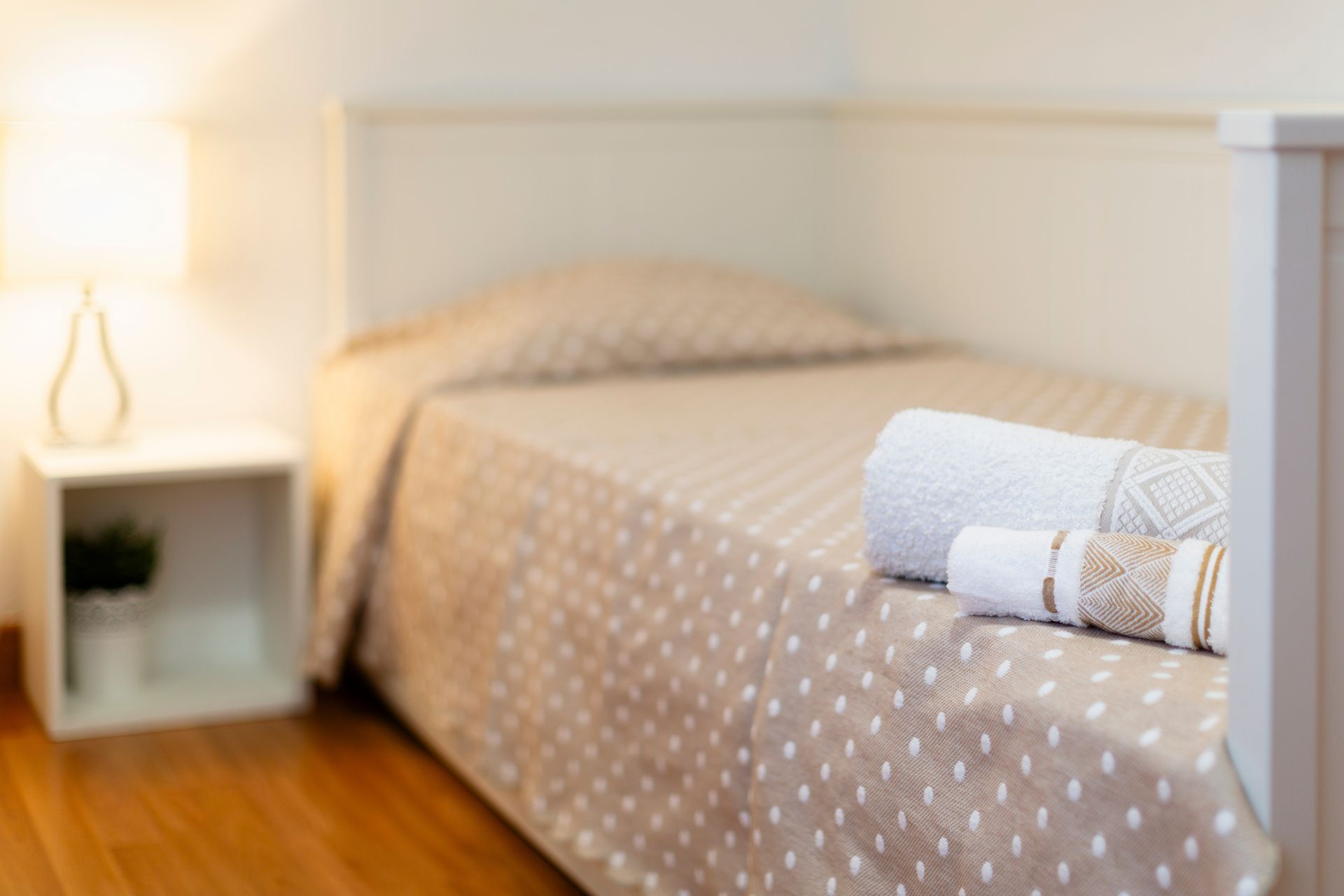 Neatly made bed with beige polka-dot cover and rolled towels in a bright bedroom