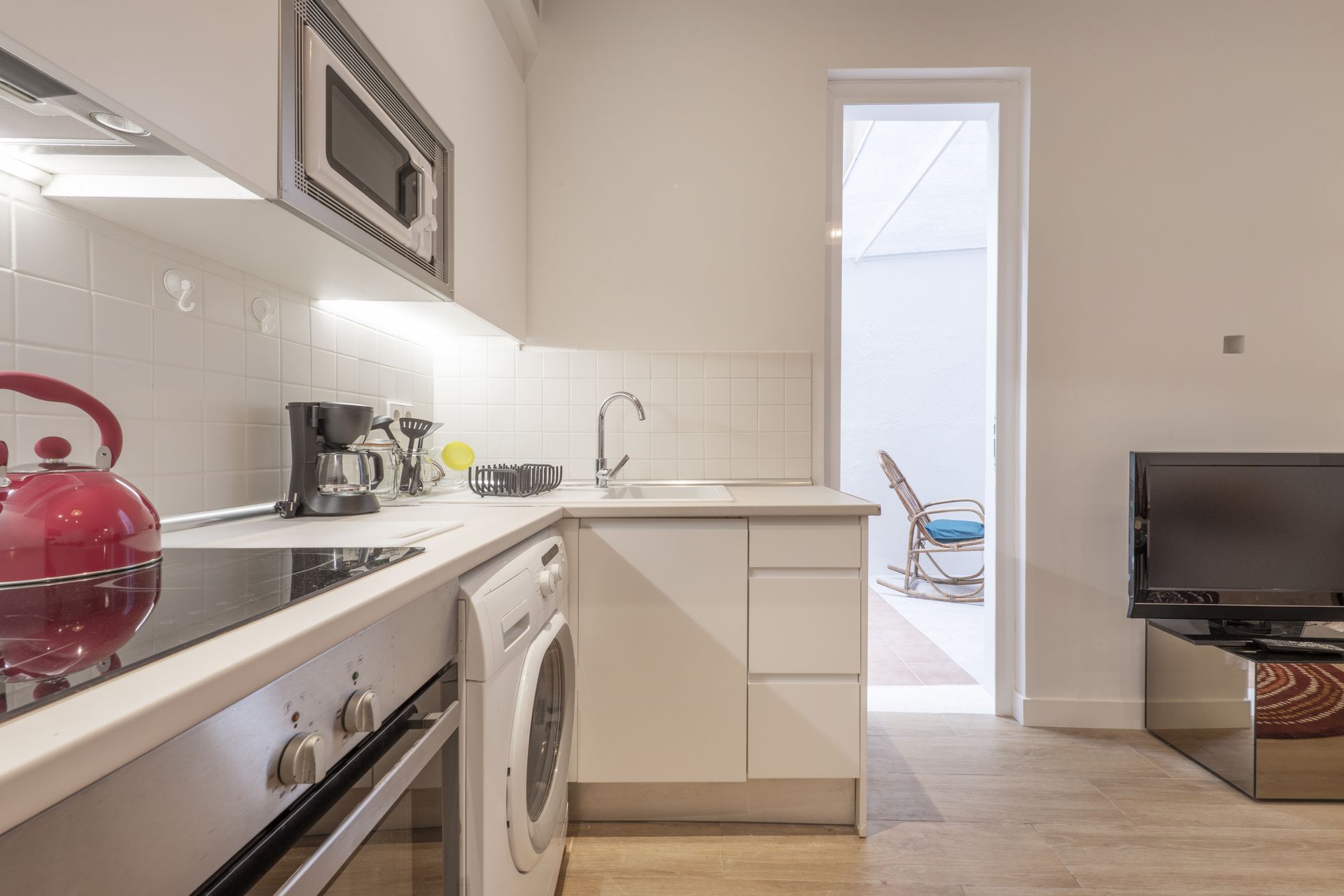 Bright kitchen with white cabinets, red kettle, washing machine, and a TV in the adjacent room.
