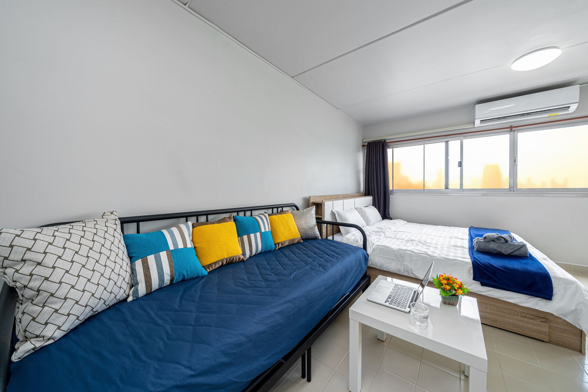 Bright modern studio with blue sofa, colorful pillows, bed, coffee table, and window air conditioner