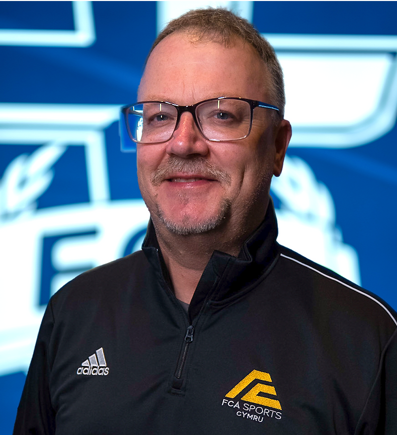 A white man in his fifties with spectacles, light hair, a light beard, and a black collared shirt with the FCA Sports Europe logo.