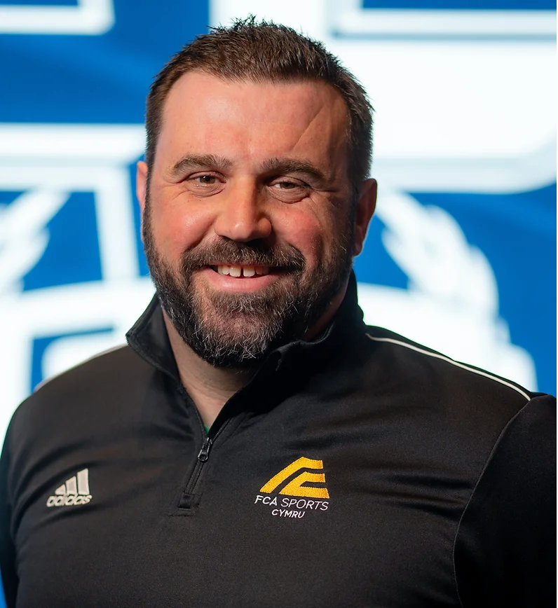 A white man in his forties with dark hair, a dark beard, and a black collared shirt with the FCA Sports Europe logo.