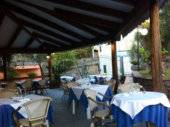 Terrazza esterna del ristorante con tovaglie blu e bianche e sedie in vimini sotto una tettoia in legno ombreggiante.