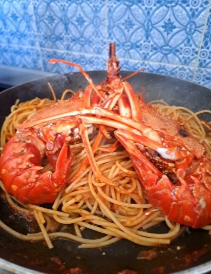 Piatto di spaghetti conditi con grossi gamberi rossi in salsa scura, su uno sfondo blu a motivi geometrici.