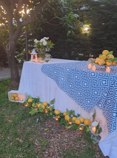 Tavolo da esterno con tovaglia bianca e blu, decorato con limoni e fiori in un contesto da giardino.