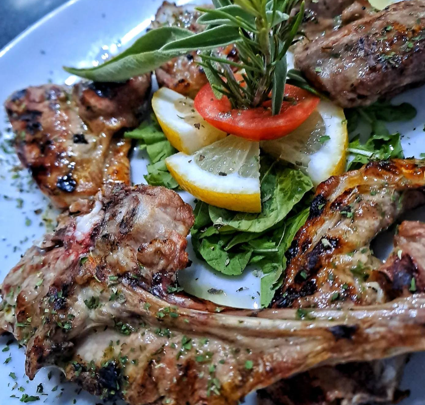 Costolette di agnello alla griglia guarnite con fette di limone, pomodoro ed erbe aromatiche, servite su un piatto bianco.
