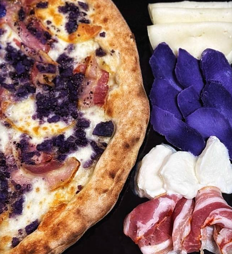 Pizza con decorazioni viola a forma di fiore accanto a fette di formaggio e salumi su sfondo scuro.